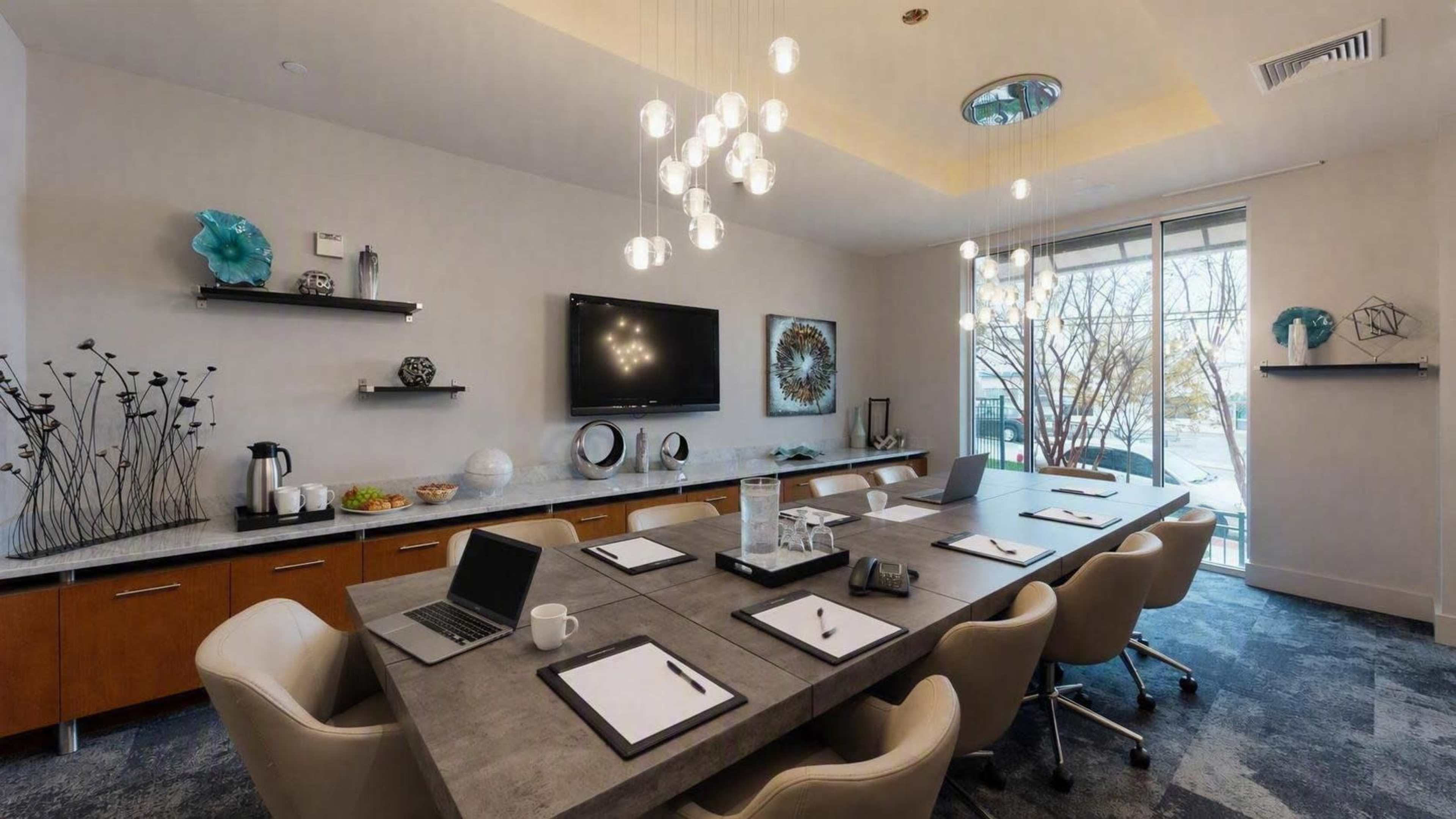 The image shows a modern conference room with a long table, comfortable chairs, and a large screen mounted on the wall, featuring a decorative arrangement on the shelves.
