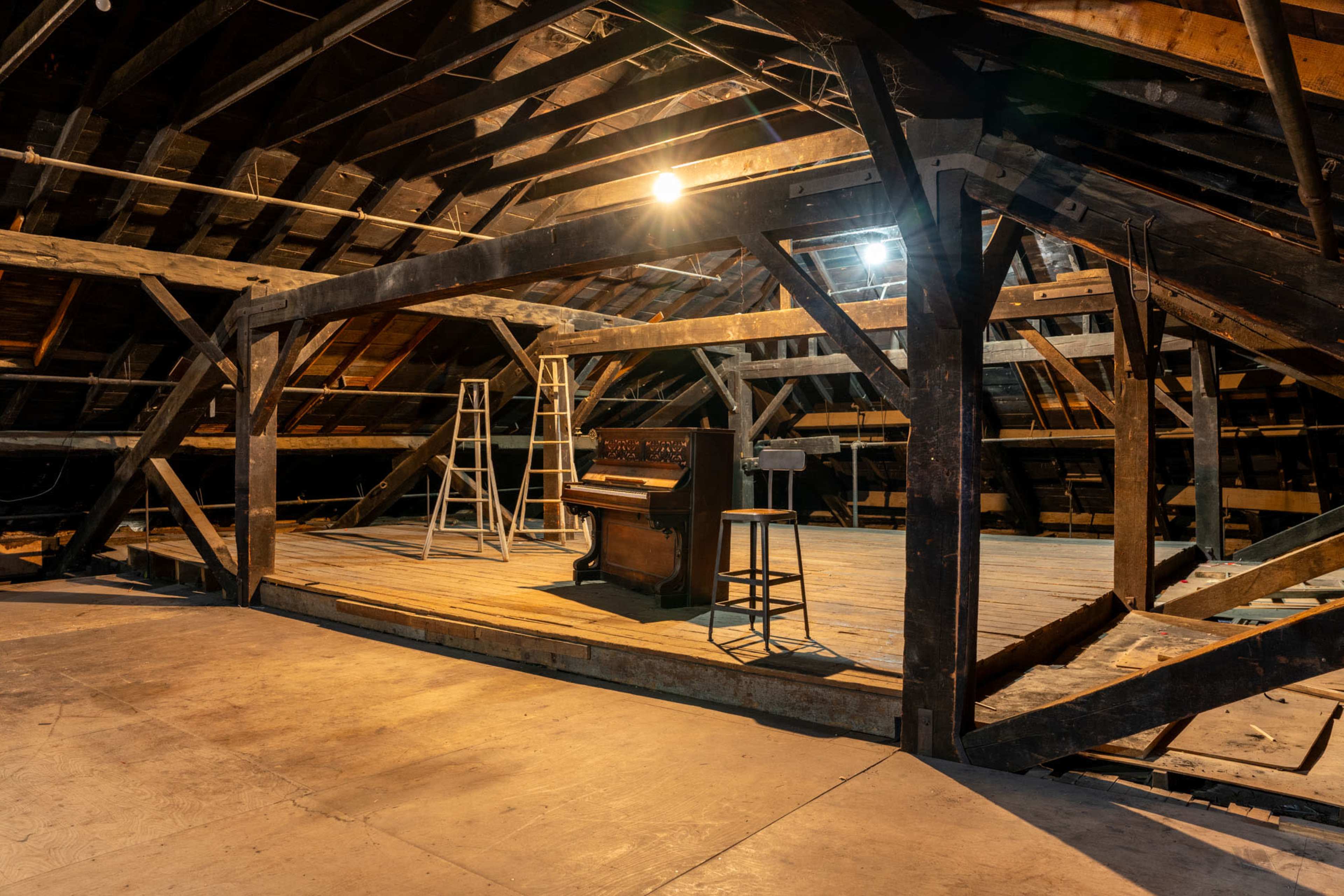 An unfinished attic with wooden beams, a piano, a stool, and two ladders positioned on the wooden floor.