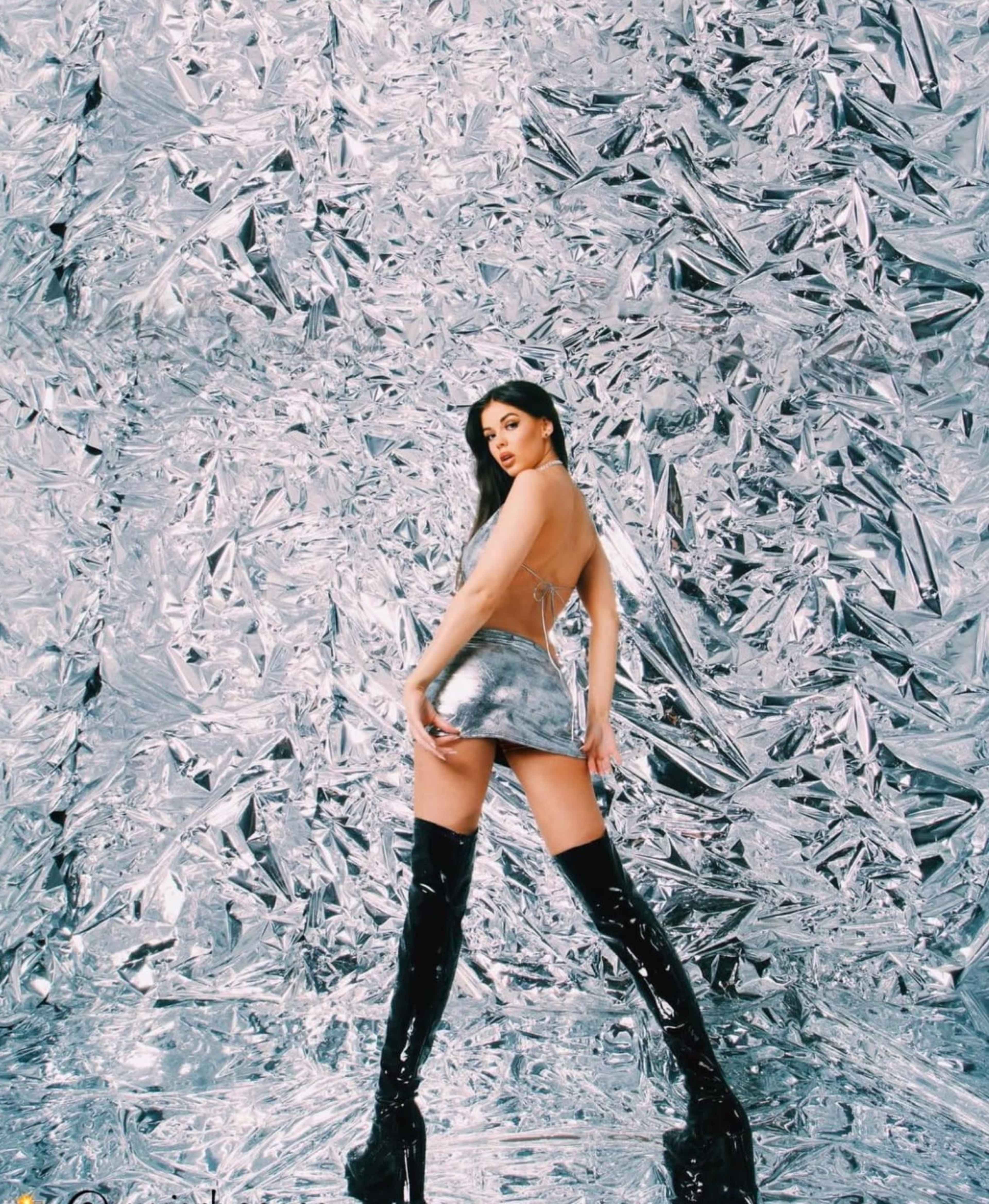 A model poses against a shimmering silver foil backdrop while wearing a metallic dress and knee-high black boots.