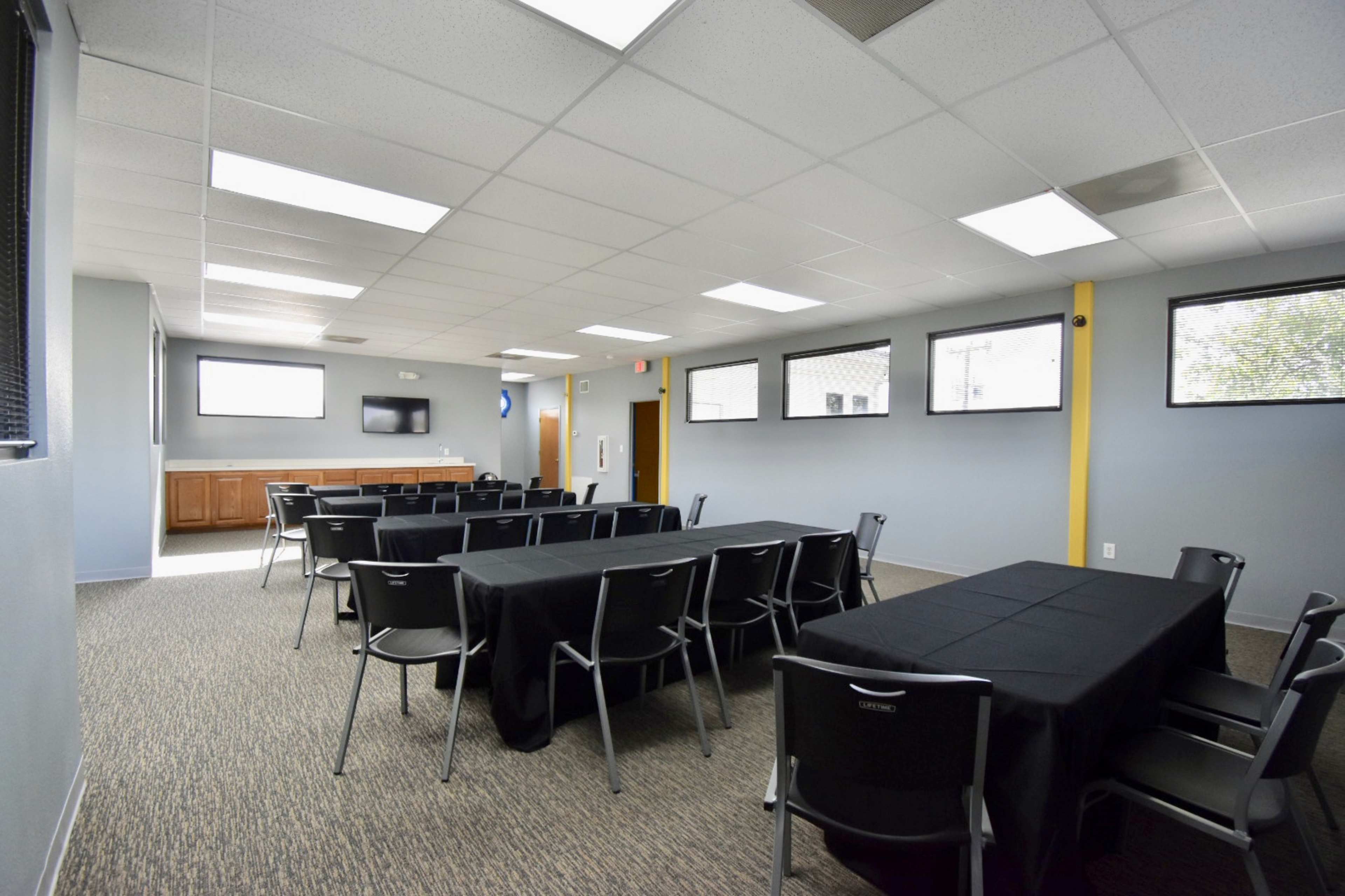 The image shows a meeting room with several long tables arranged in rows, black tablecloths, and chairs, with windows allowing natural light.