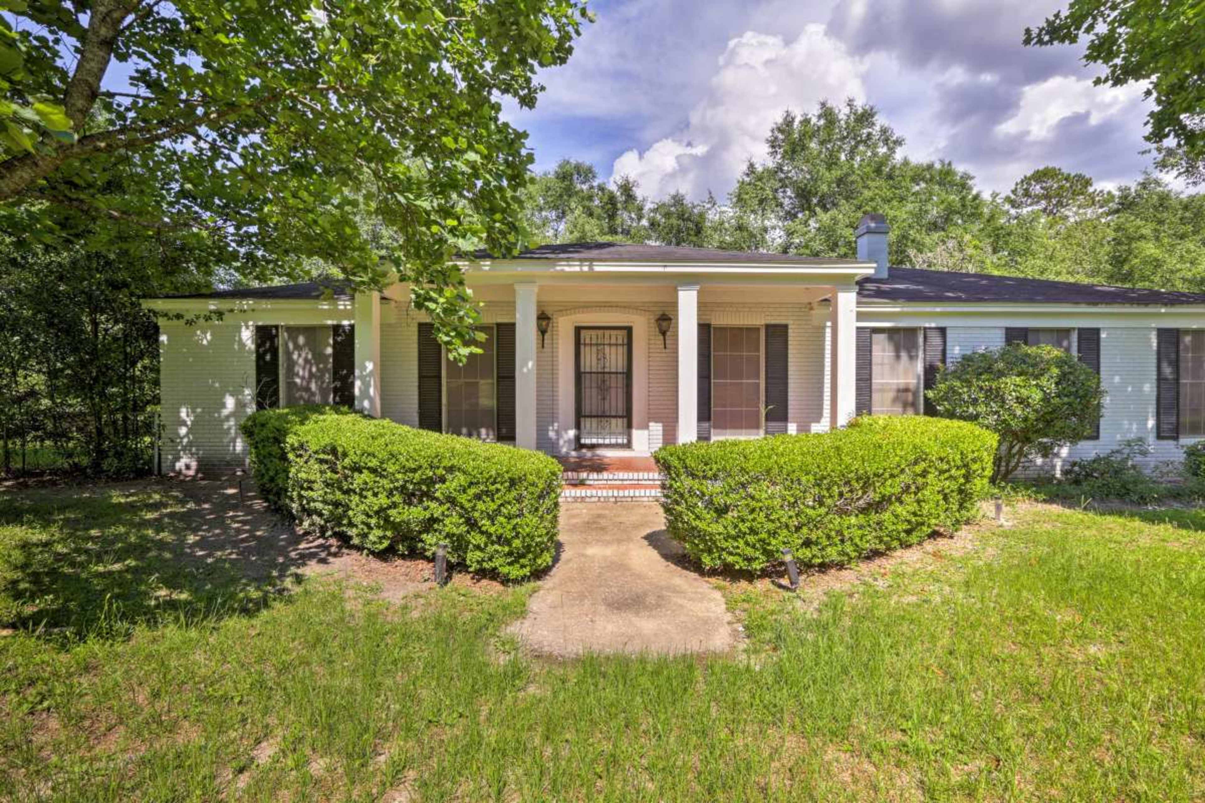 A single-story house with a front porch is flanked by well-trimmed bushes and a grassy lawn under a blue sky with clouds.