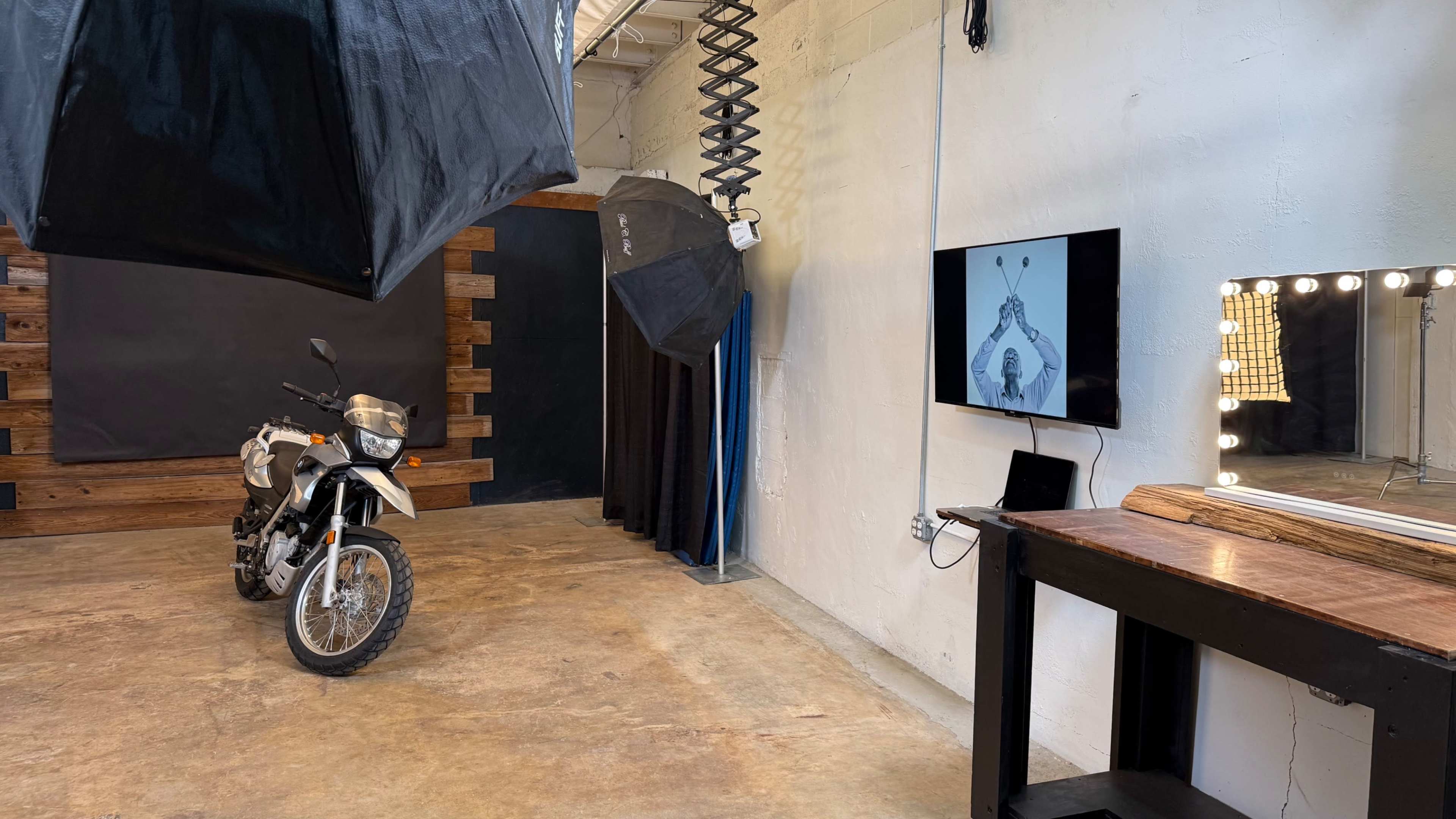 The image shows a photography studio featuring a motorcycle, a large black backdrop, soft boxes, and a television displaying an image.