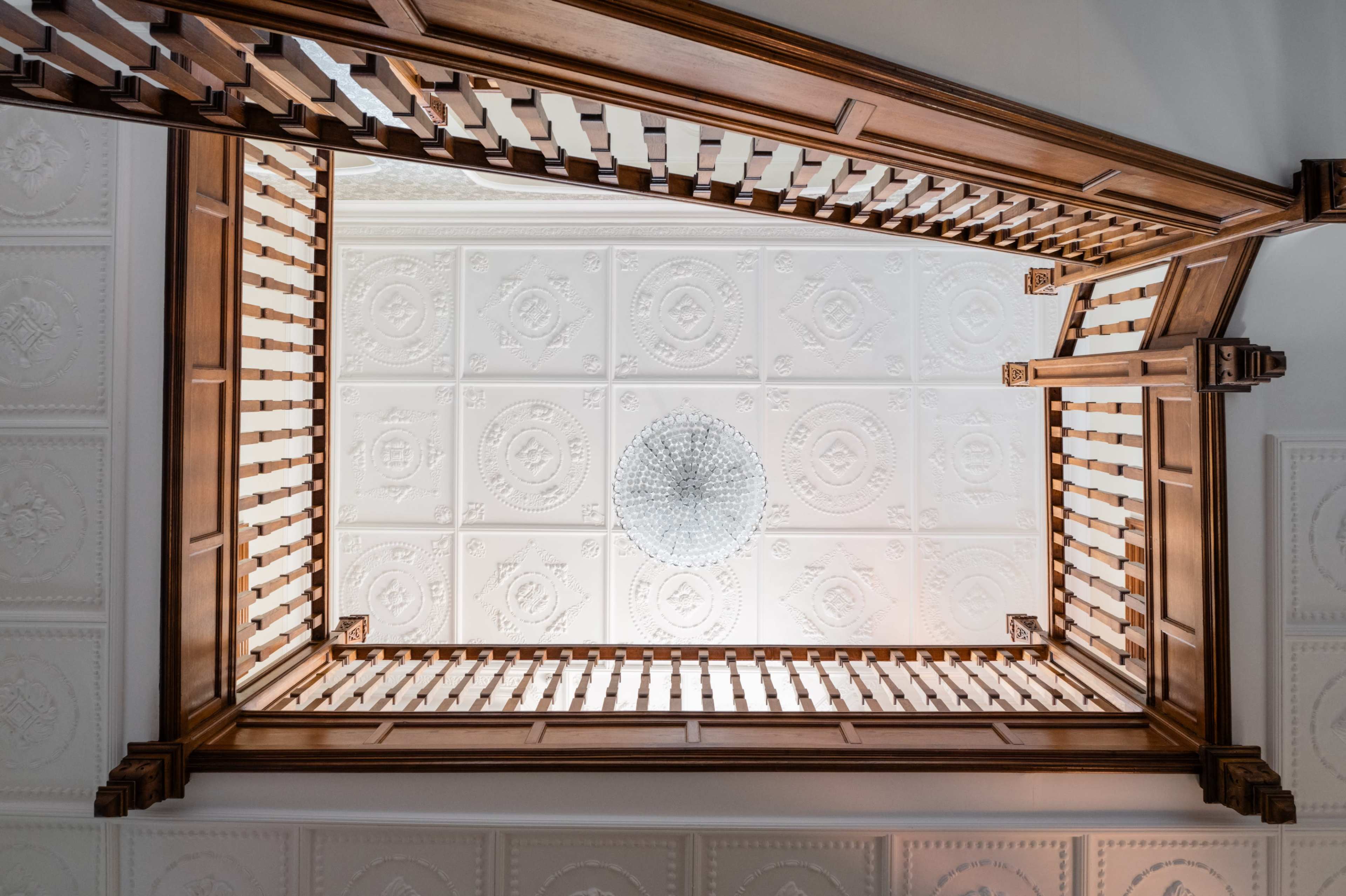 A wooden staircase viewed from below, leading to an ornate ceiling with a chandelier at the center.