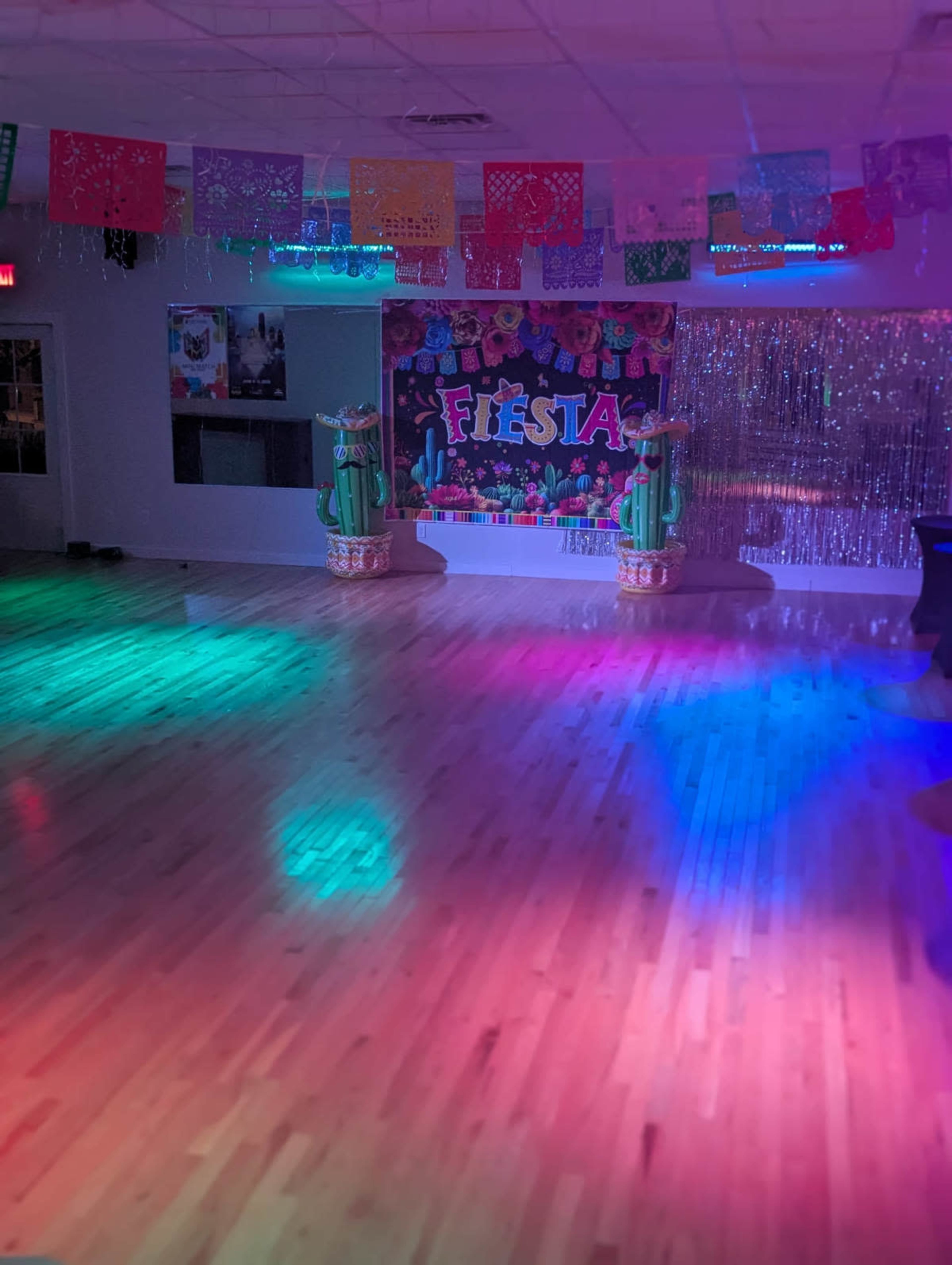 The image shows a decorated dance floor illuminated by colorful lights, featuring a "Fiesta" backdrop and floral decorations.