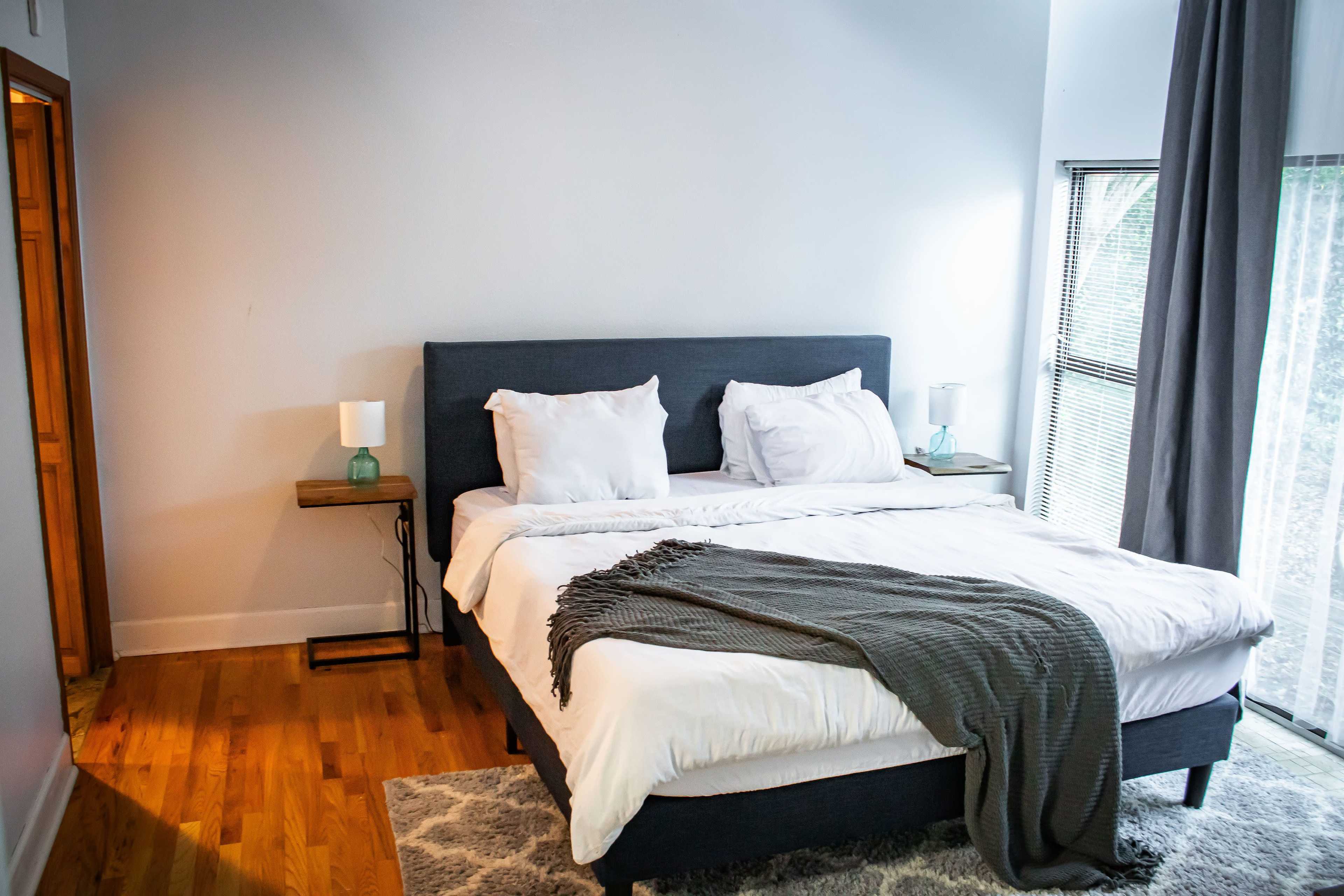 A neatly arranged bedroom featuring a bed with white bedding, a gray blanket, and two side tables with lamps beside it, along with a window letting in natural light.