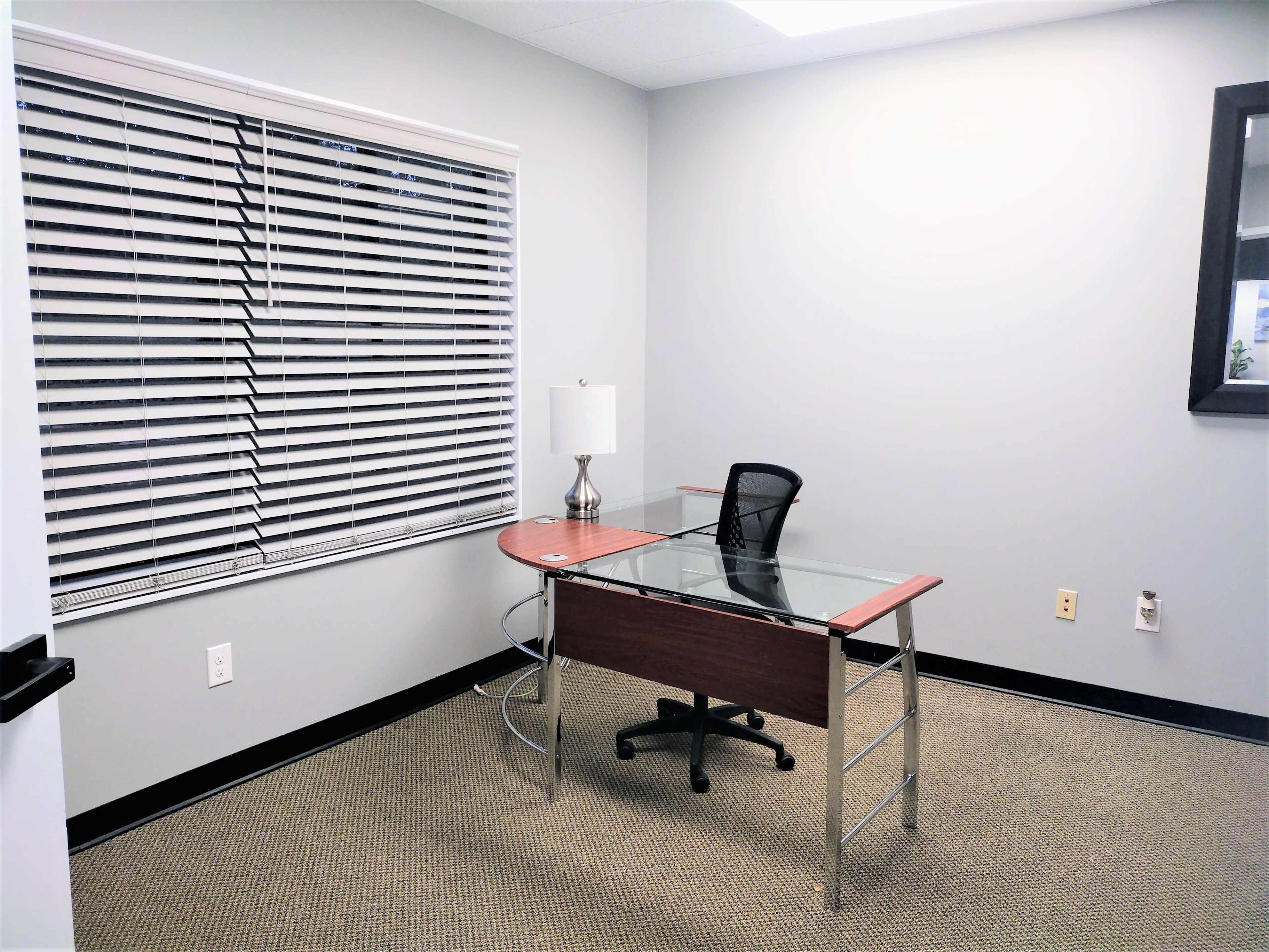 A sparsely furnished office space with a glass-top desk, a black rolling chair, and a lamp beside the window.