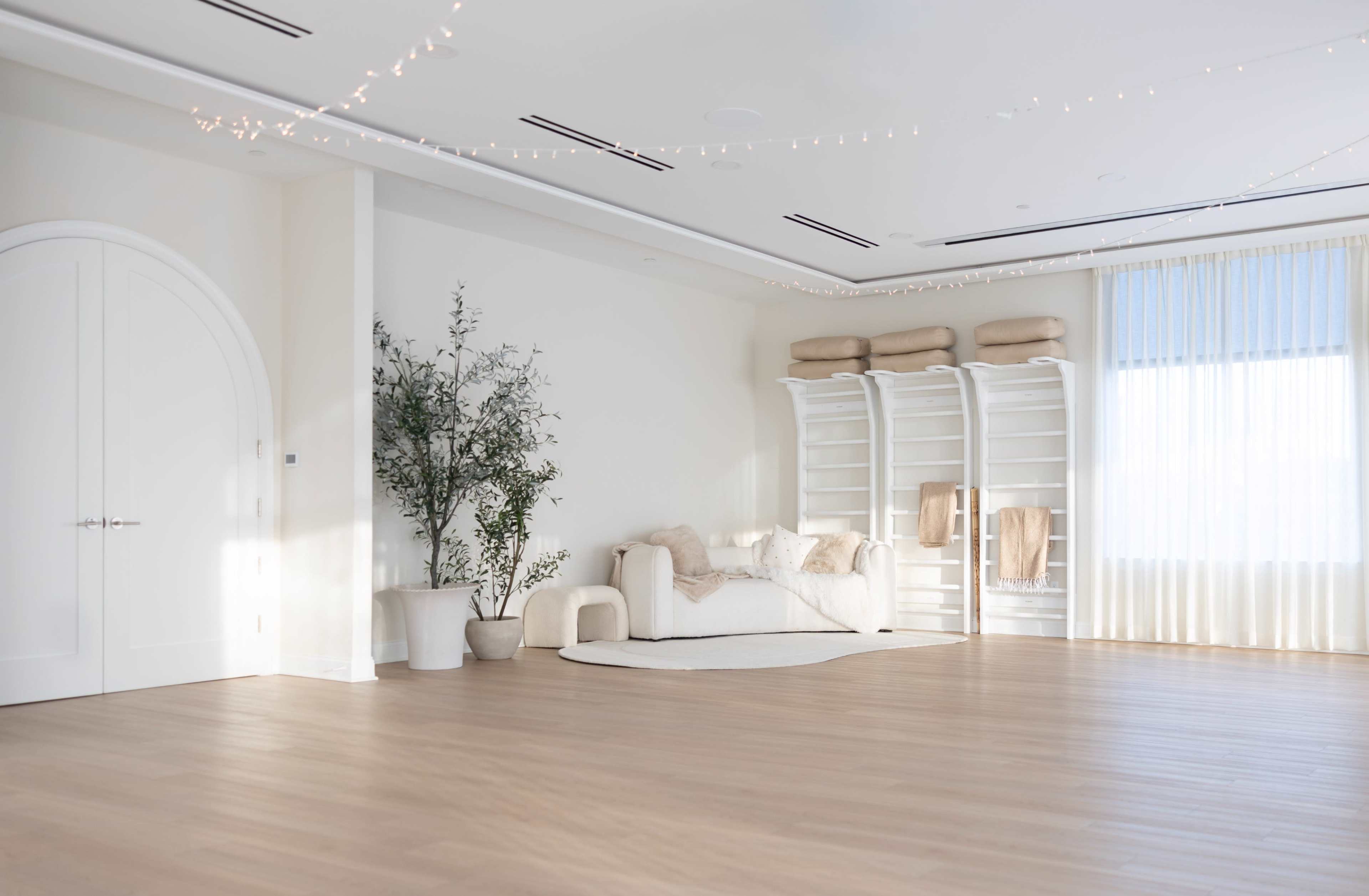 The image shows a spacious, softly lit room with a white couch, towels hanging on a shelf, and a potted plant.