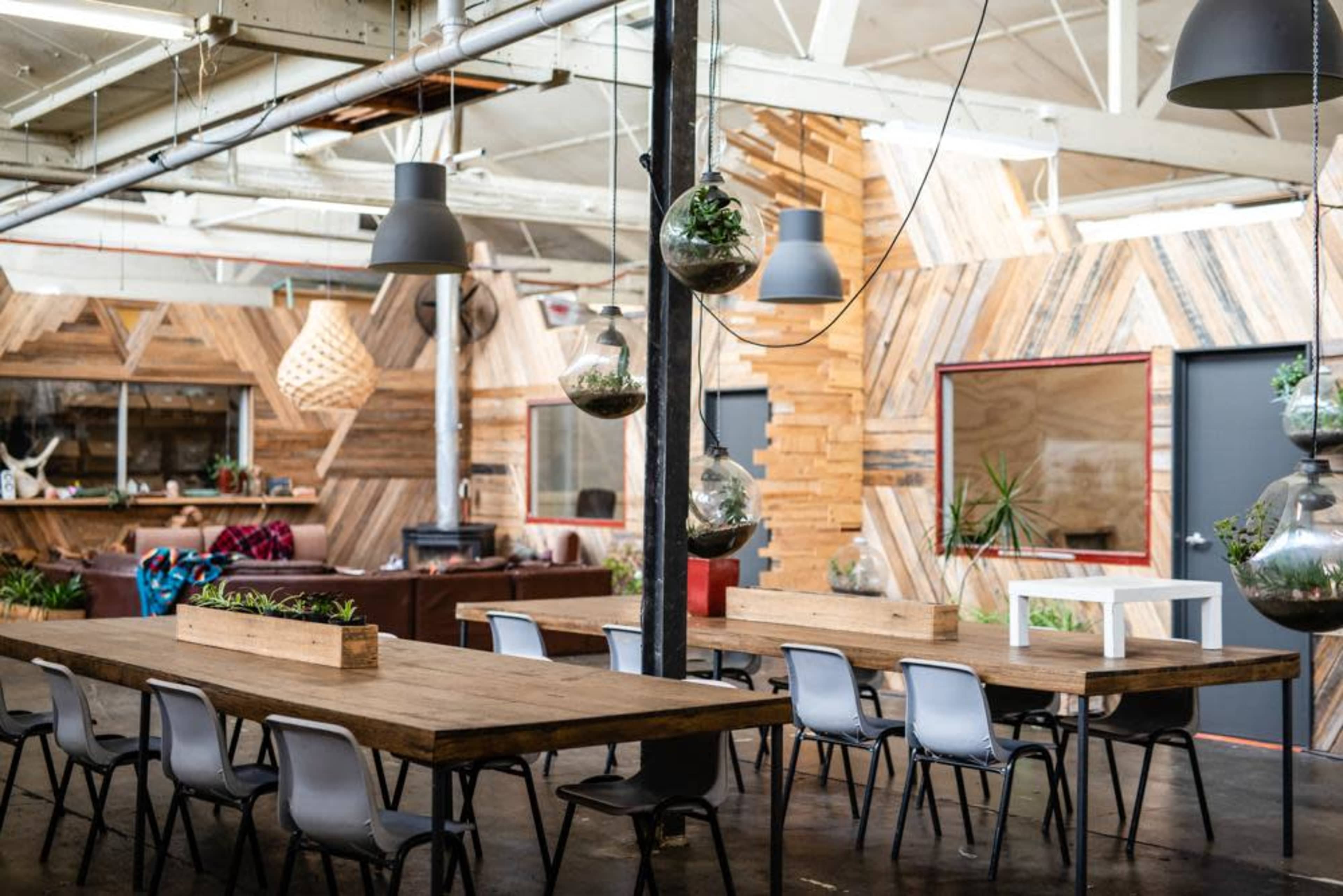 A spacious, modern interior with wooden decor, featuring long tables, hanging planters, and industrial lighting.