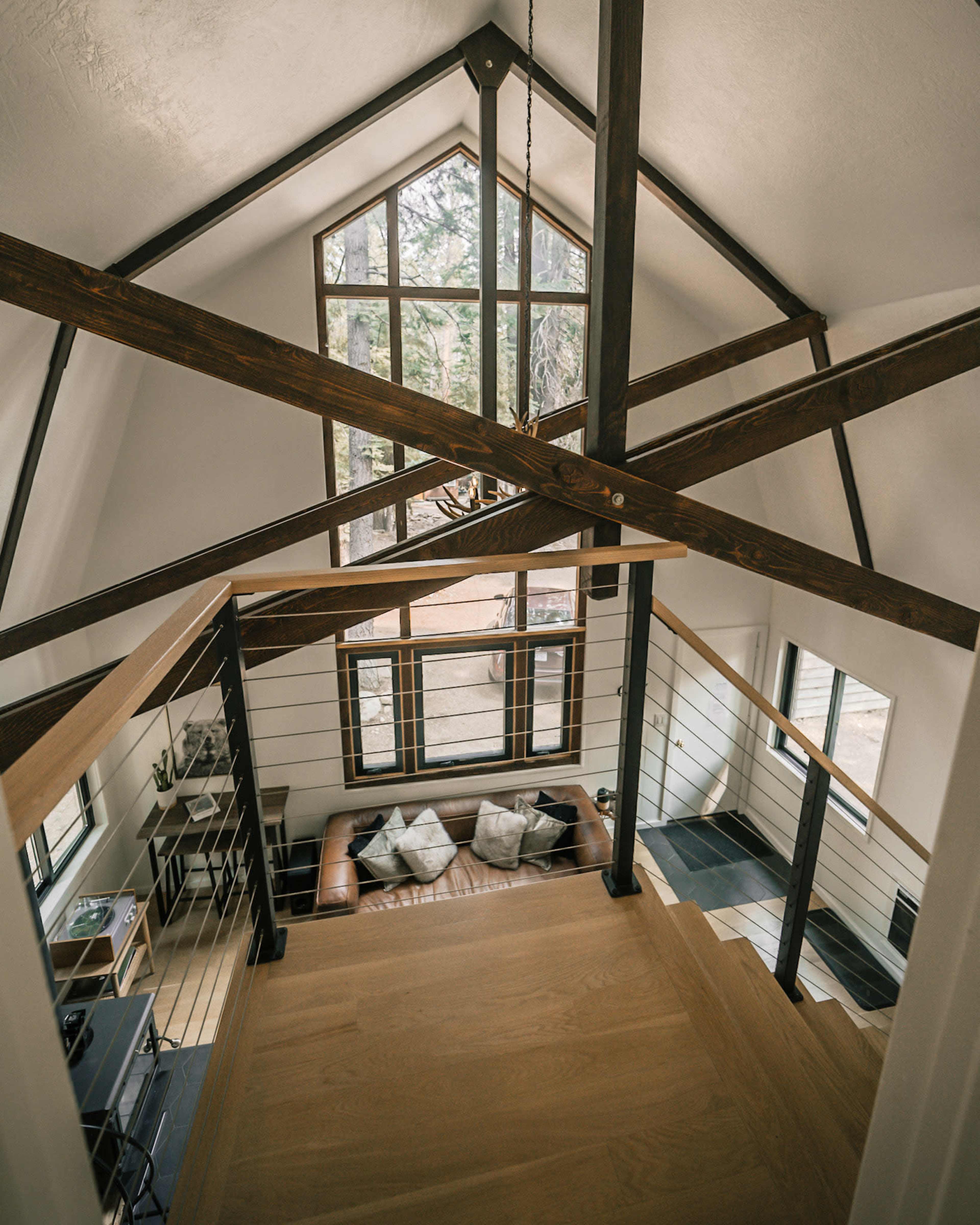 A spacious two-story interior with a wooden staircase, large windows, and exposed beams, overlooking a living area with a couch and pillows.