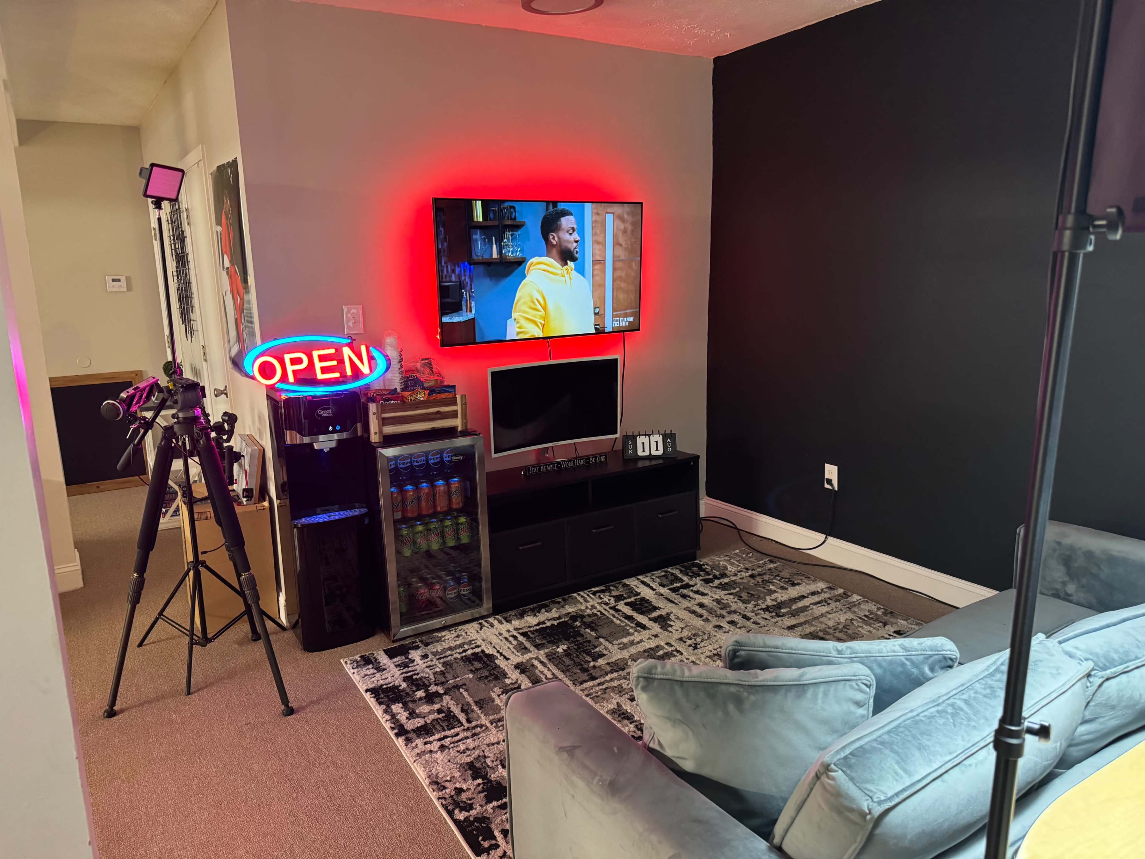 A living room setup with a large TV displaying a show, a neon "OPEN" sign, a camera on a tripod, and a shelf with various items, all against a dark wall.