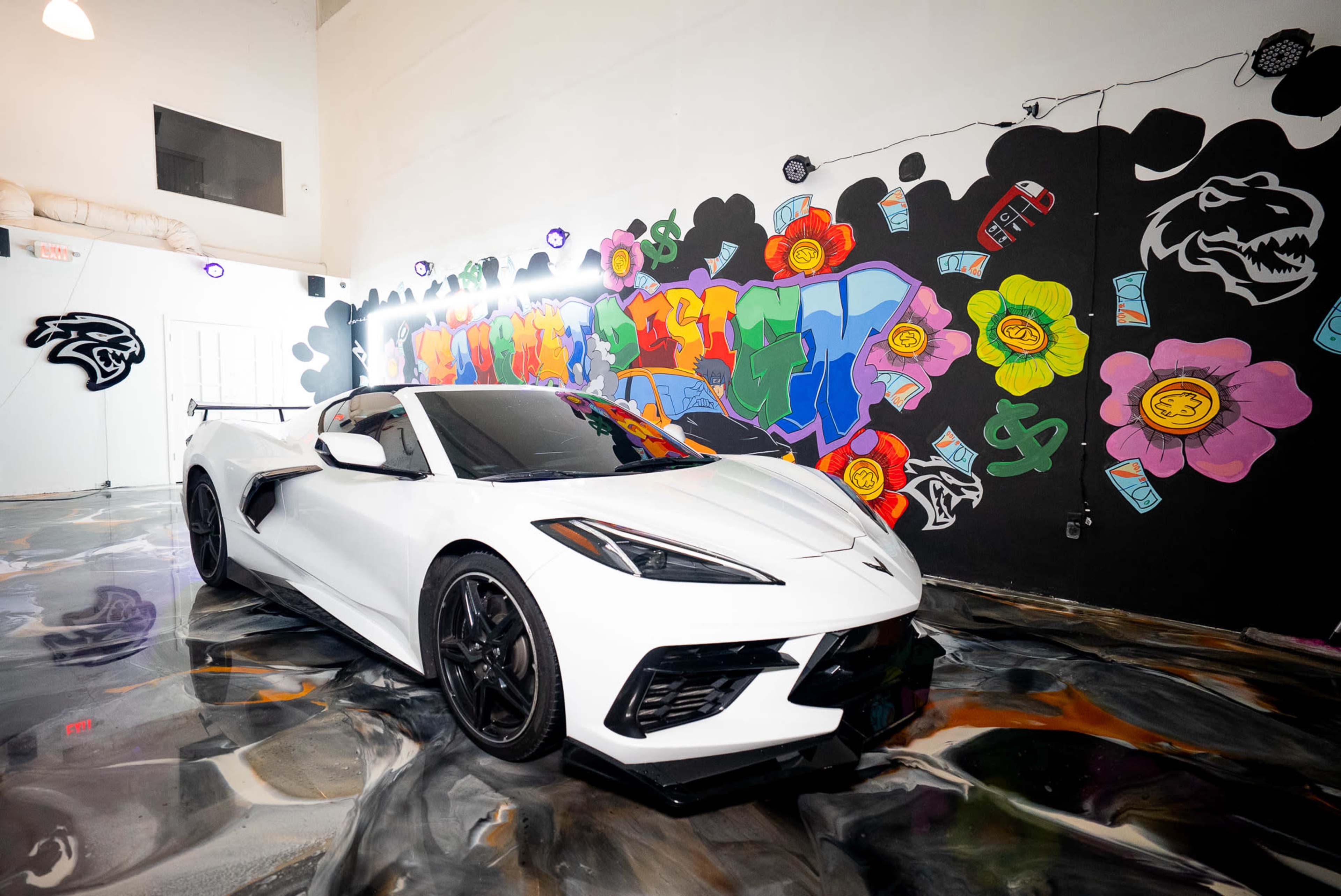 A white Corvette C8 is parked on a colorful, graffiti-decorated floor in an indoor space.