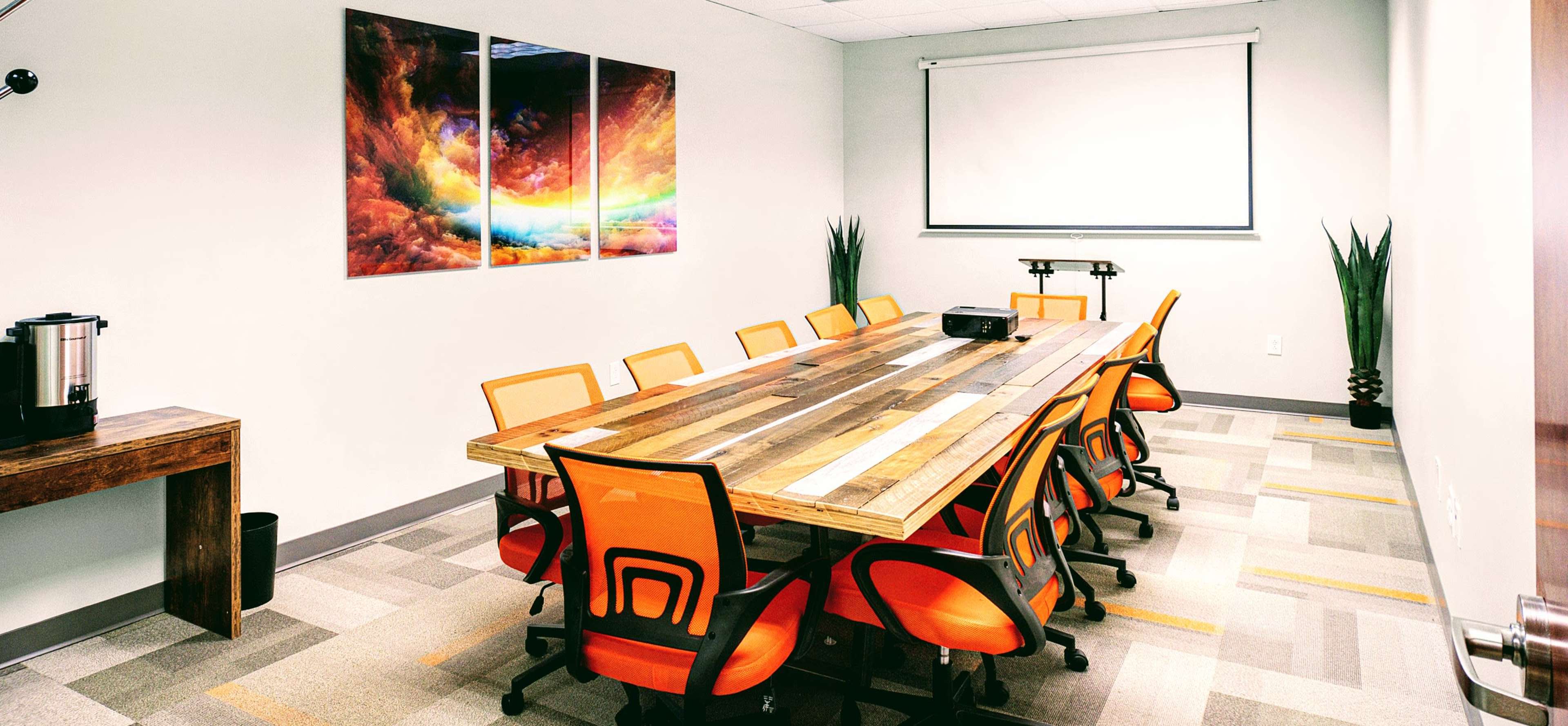 A conference room features a large wooden table surrounded by orange chairs, with a projector and a screen at one end, and abstract artwork on the walls.