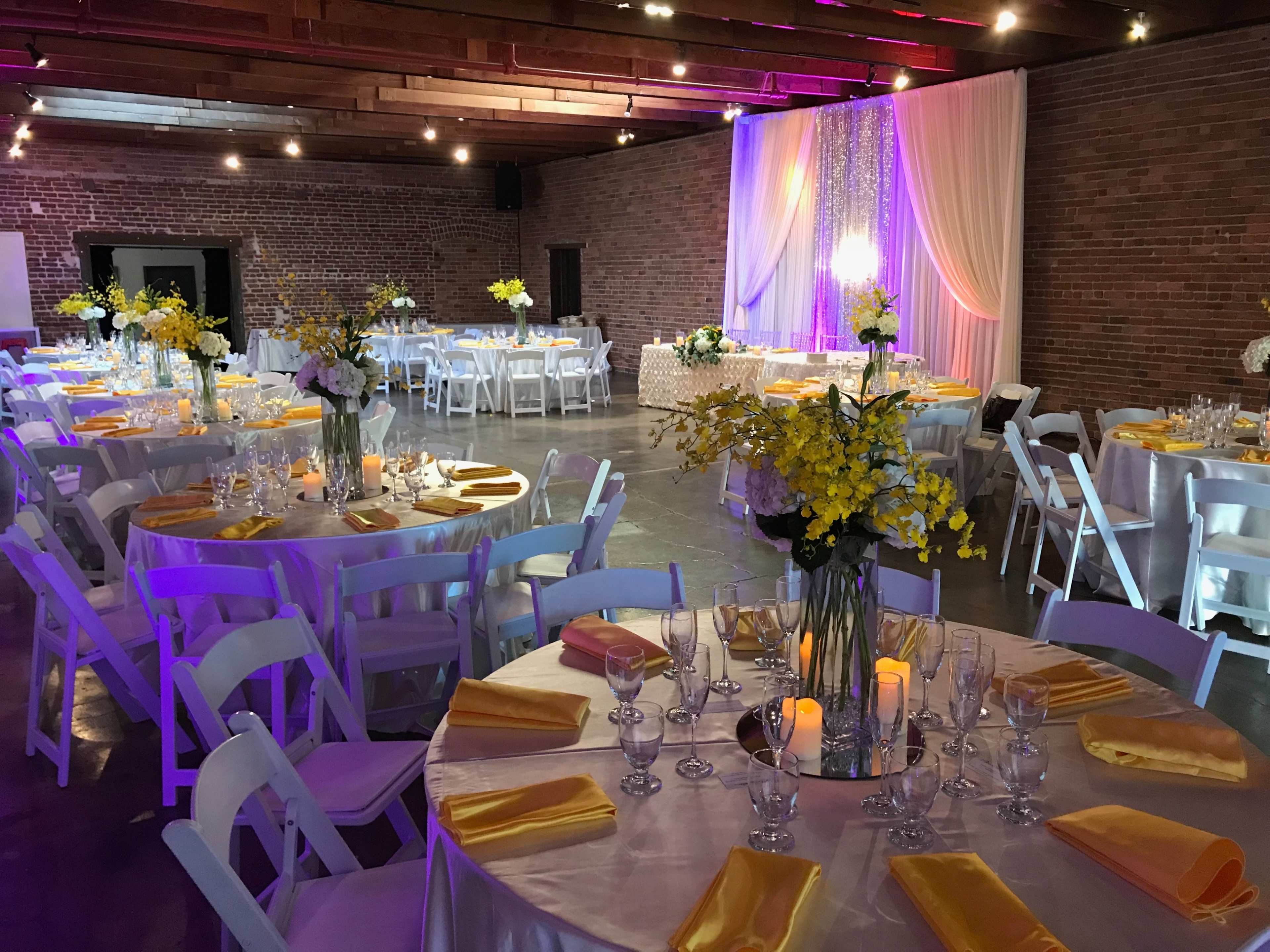 The venue features elegantly set round tables with yellow napkins and floral centerpieces, under soft lighting and draped fabric against a brick wall.