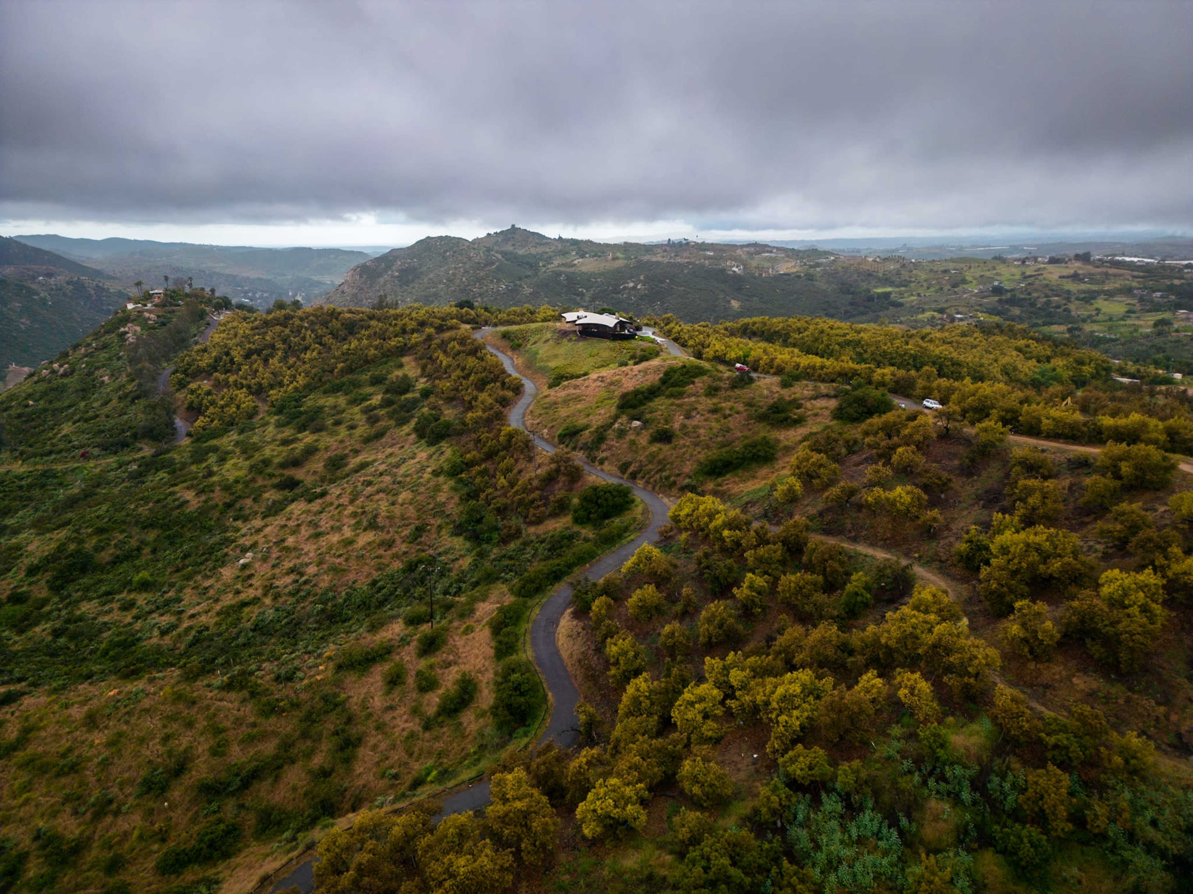 A winding road leads to a house on a hill surrounded by green vegetation and rolling hills under a cloudy sky.