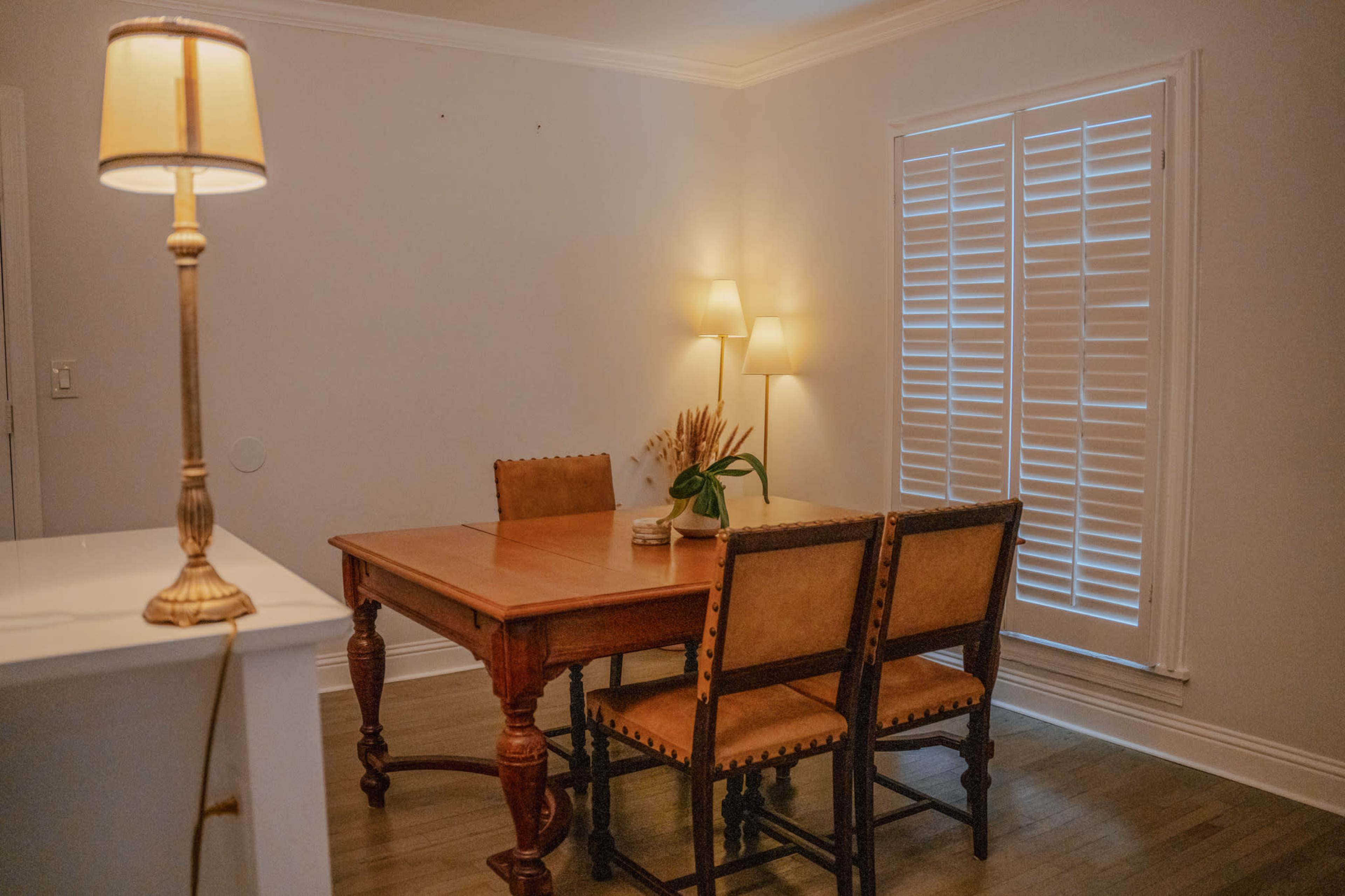 A wooden dining table with four chairs is positioned in a well-lit room featuring a lamp and window shutters.