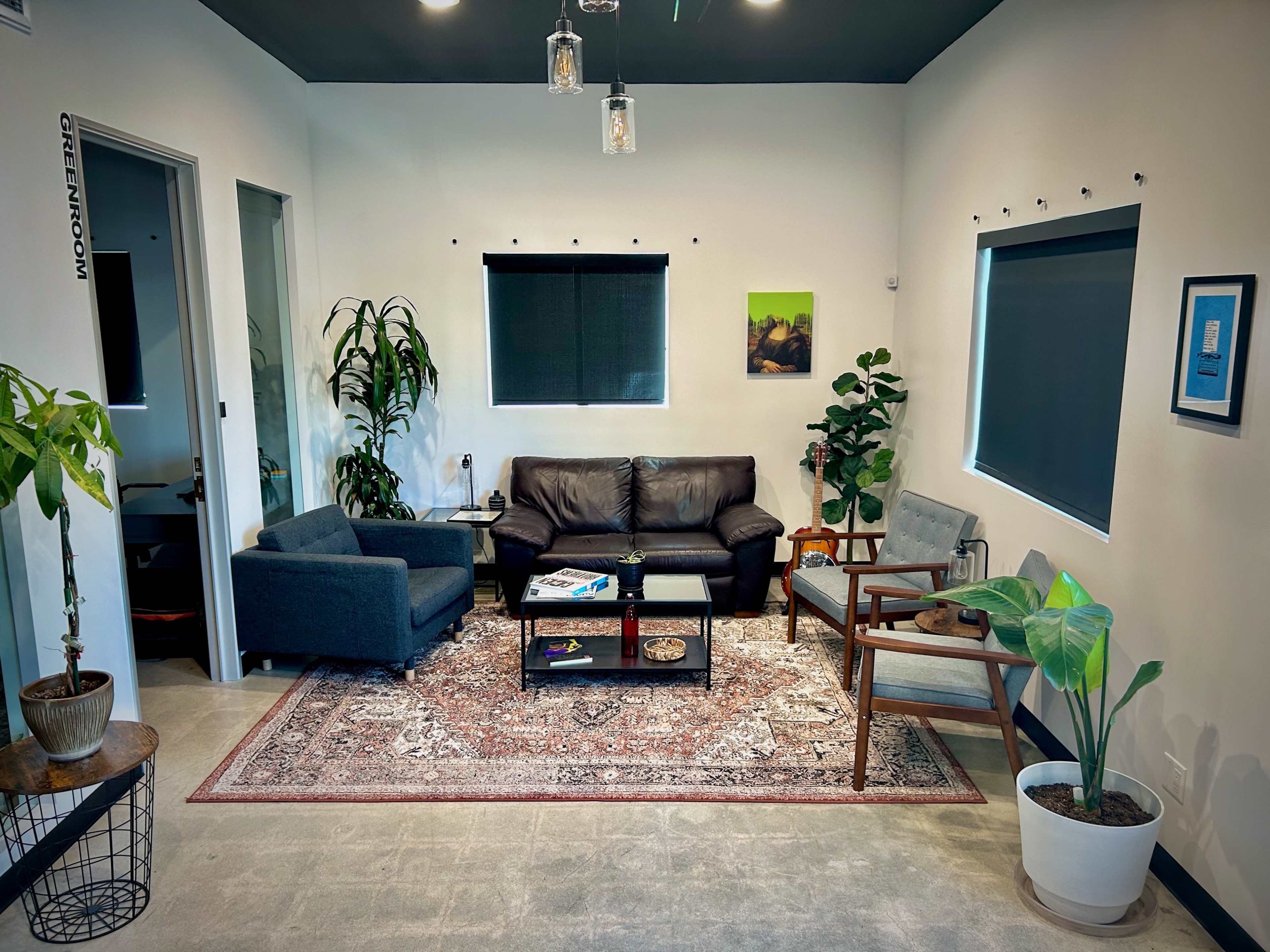 A modern lounge area features two seating arrangements, a coffee table with books, and decorative plants against a backdrop of neutral-colored walls and dark window treatments.