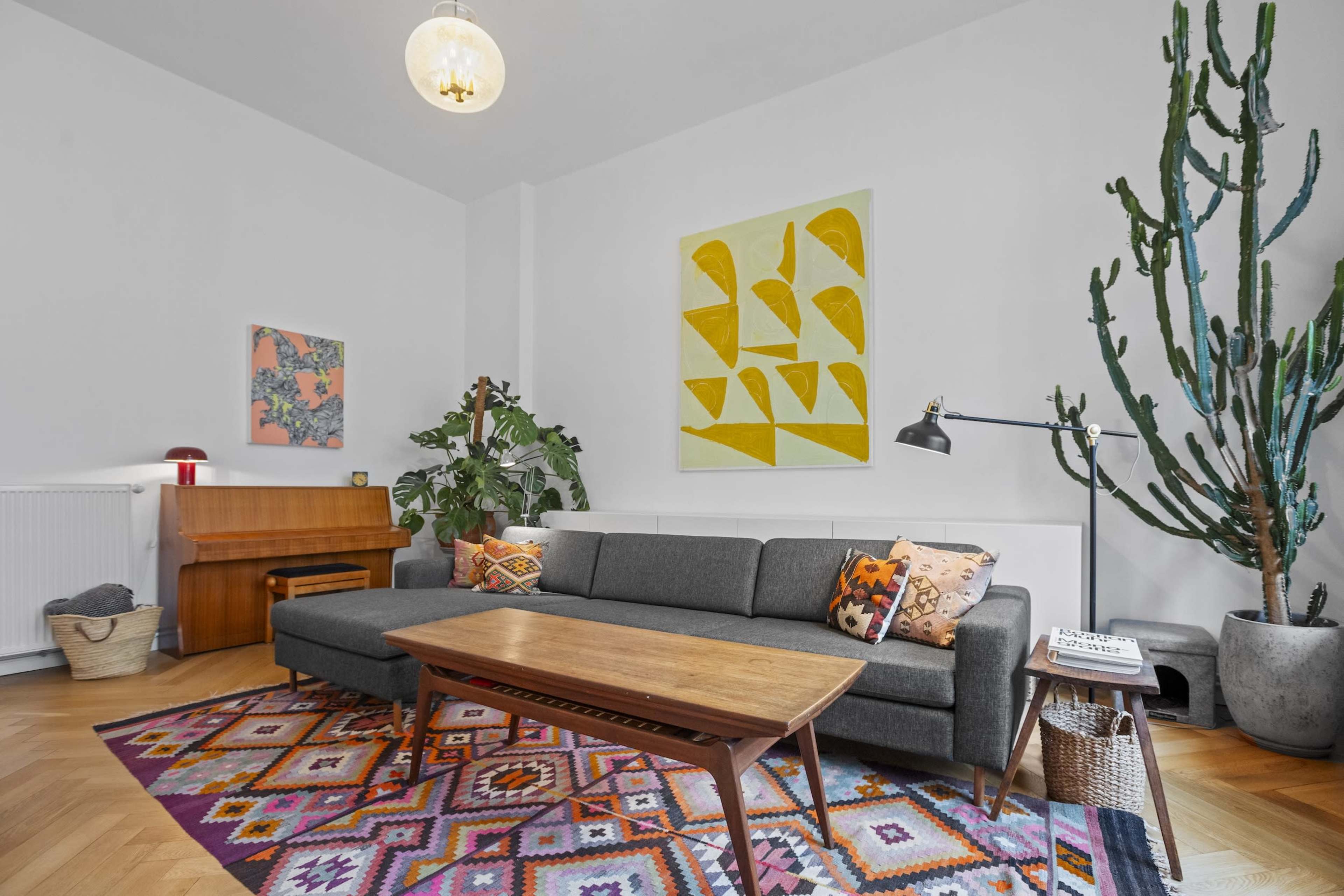 The living room features a gray sofa with colorful cushions, a wooden coffee table, a patterned area rug, and potted plants, complemented by abstract art on the walls.