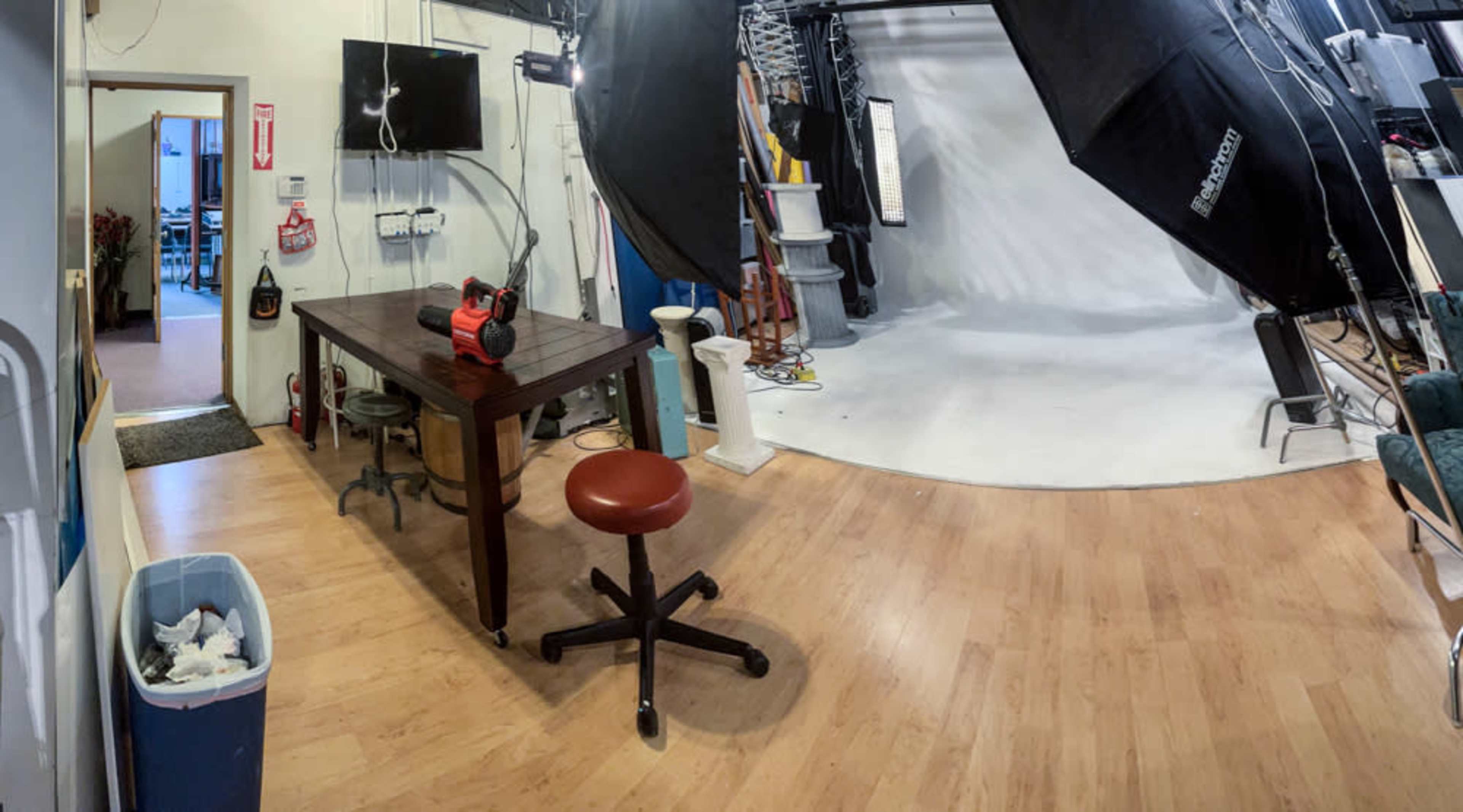 The image shows a photography studio with a wooden table, a rolling stool, and various lighting equipment set up against a plain backdrop.