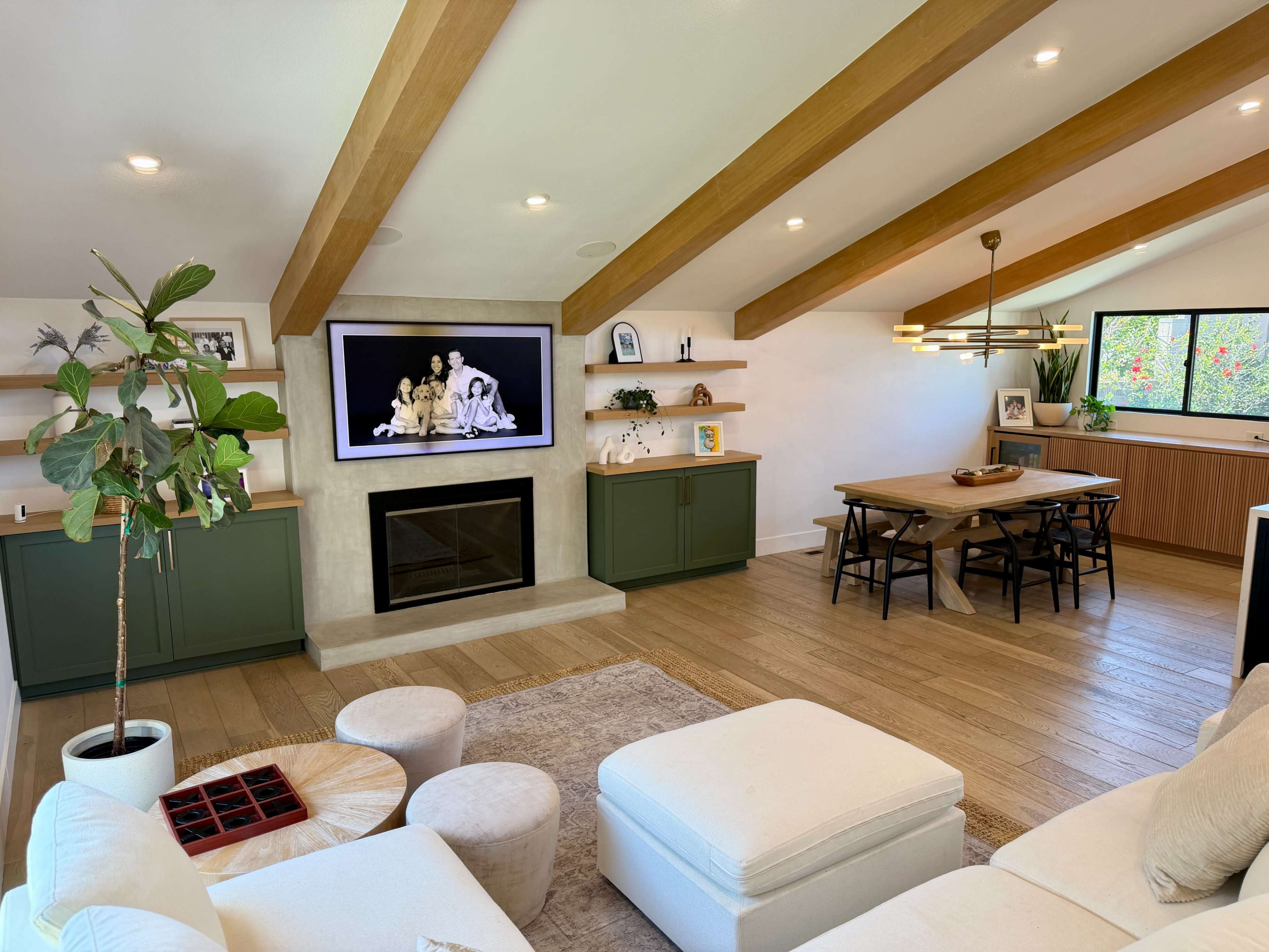 The image shows a modern living room with a large television mounted on the wall, a light-colored sectional sofa, a wooden dining table, and greenery in the corner.
