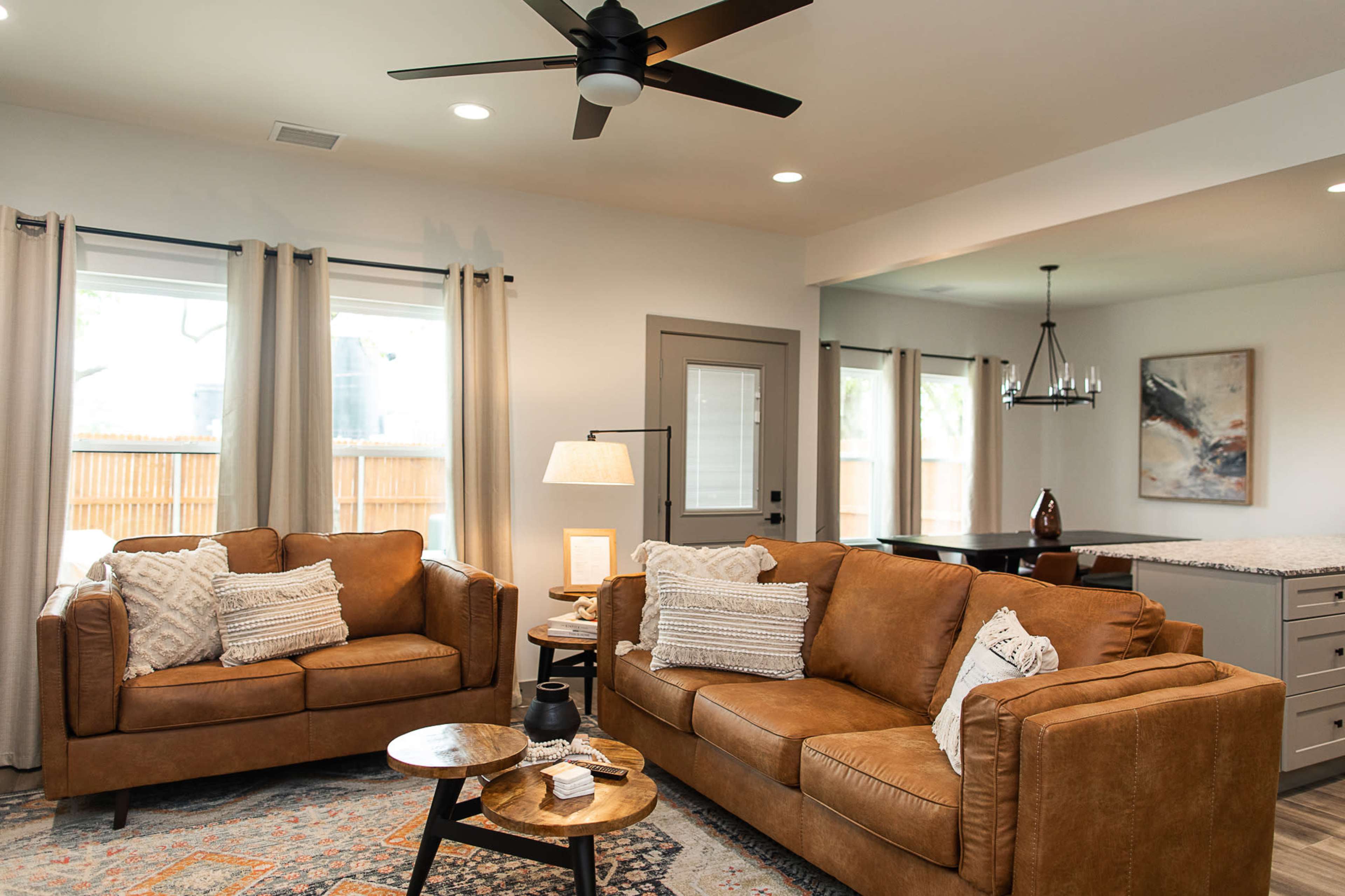 The living room features two brown leather sofas, a wooden side table, and light-colored curtains, with a dining area visible in the background.