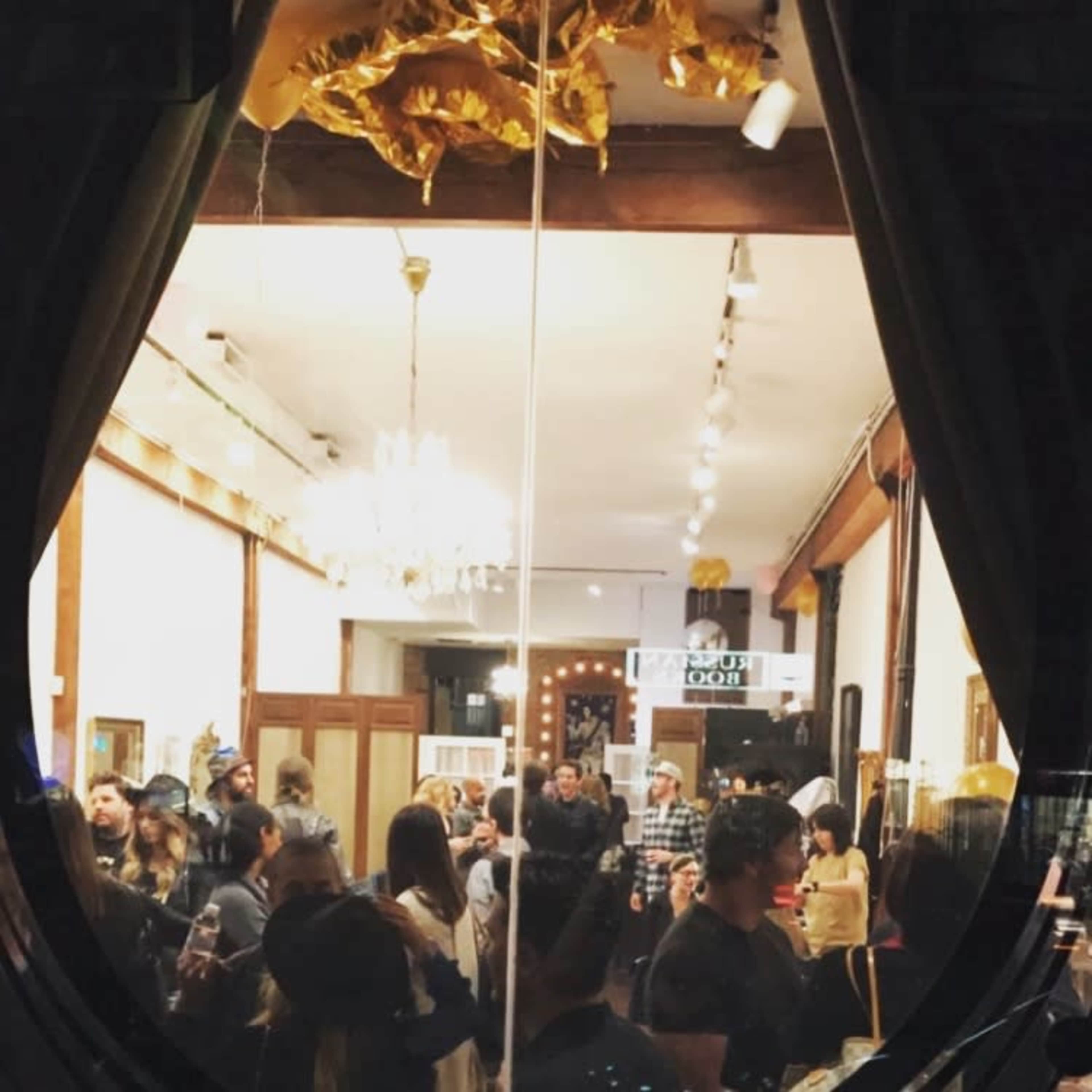 The image shows a crowded indoor gathering with people mingling under decorative lights and a chandelier.