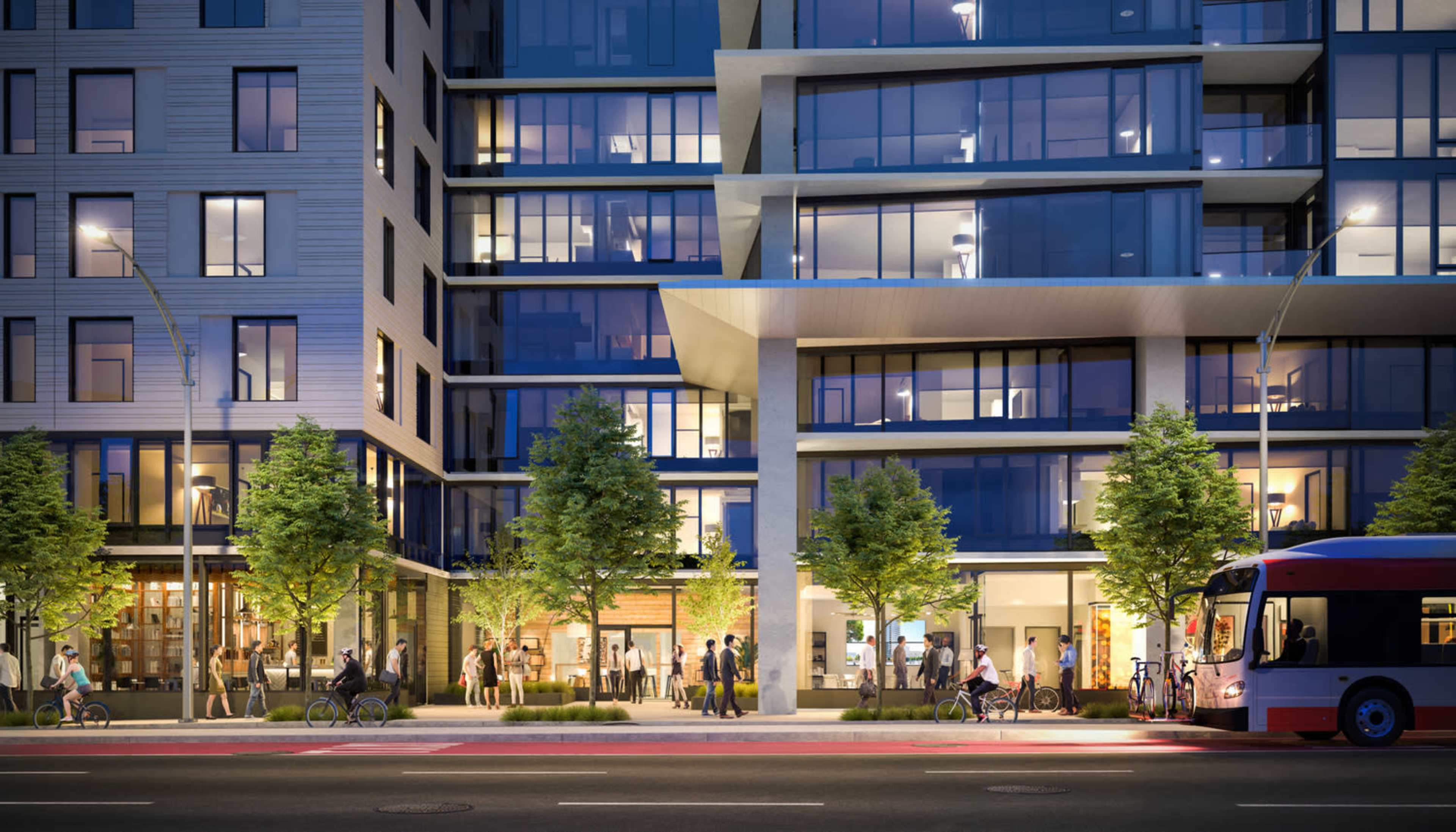 A modern multi-story building features large glass windows, storefronts at street level, and people walking and biking along the sidewalk in an urban setting.