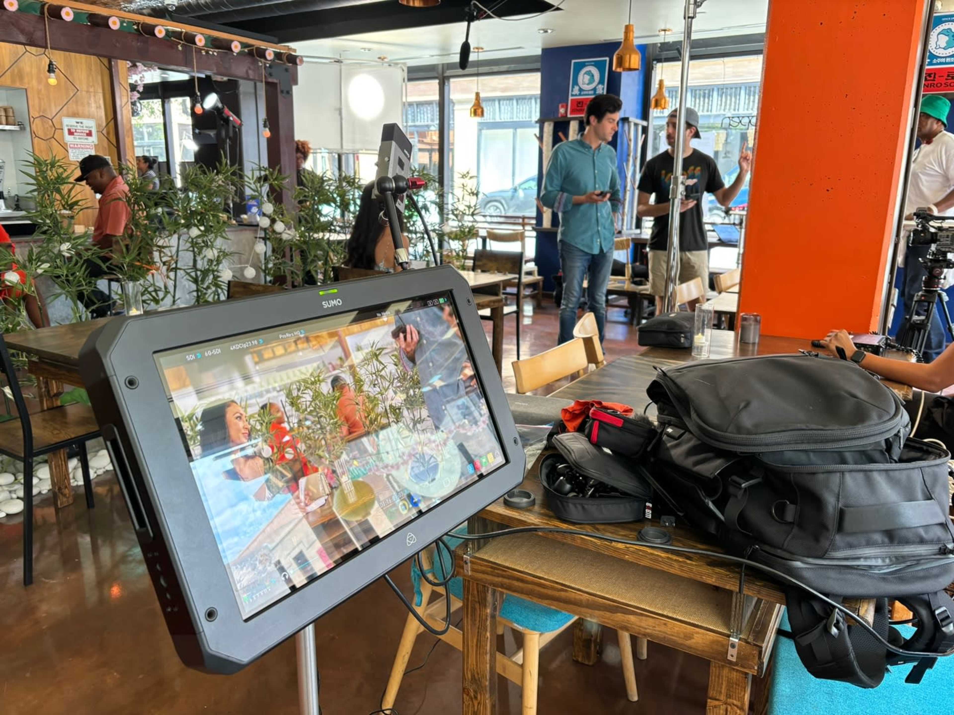 A digital monitor displays a video feed while crew members set up equipment in a café setting.