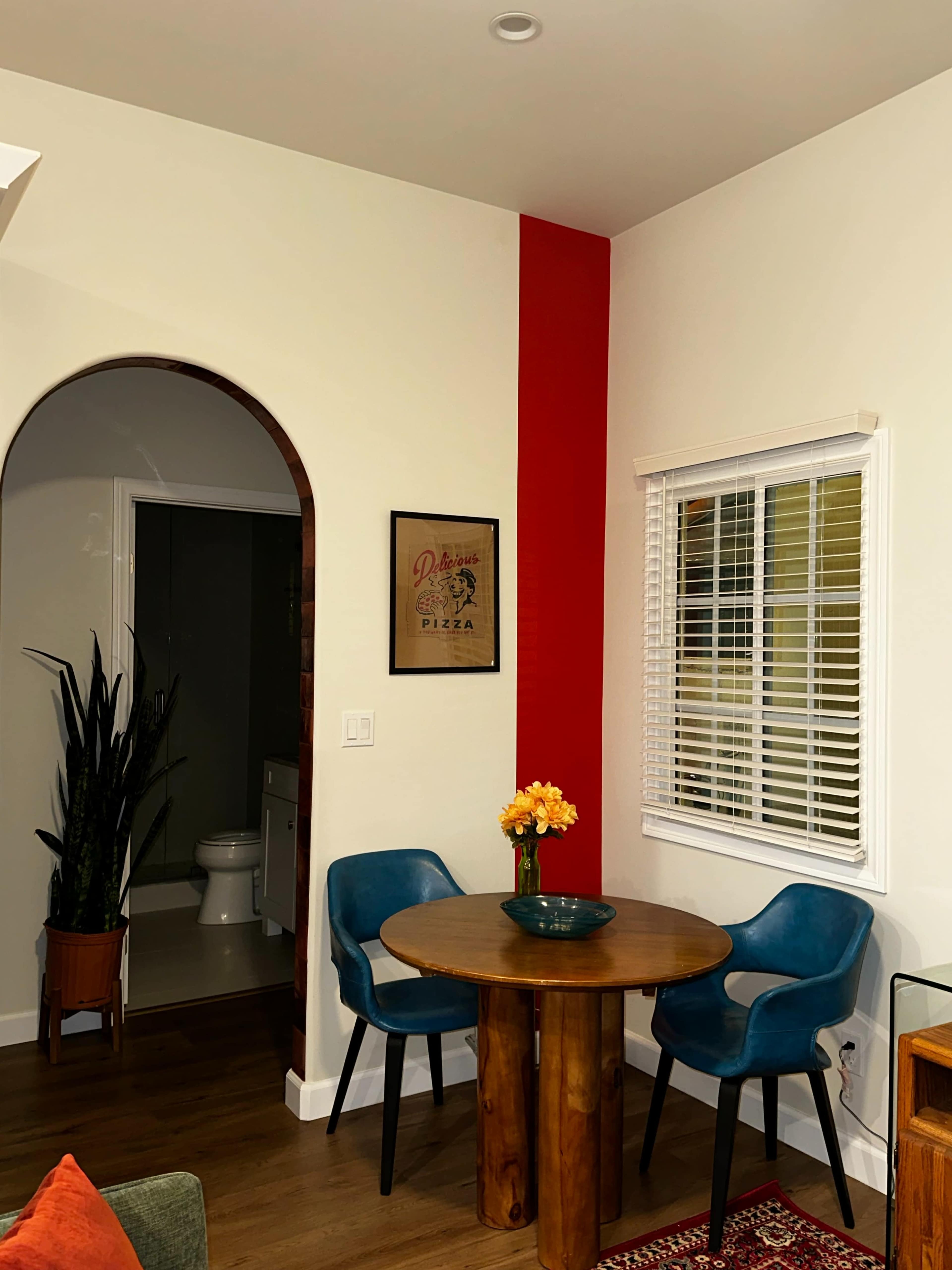 A small dining area features a round wooden table with two blue chairs, potted plants in the corner, and a framed decorative piece on the wall beside a red accent section.