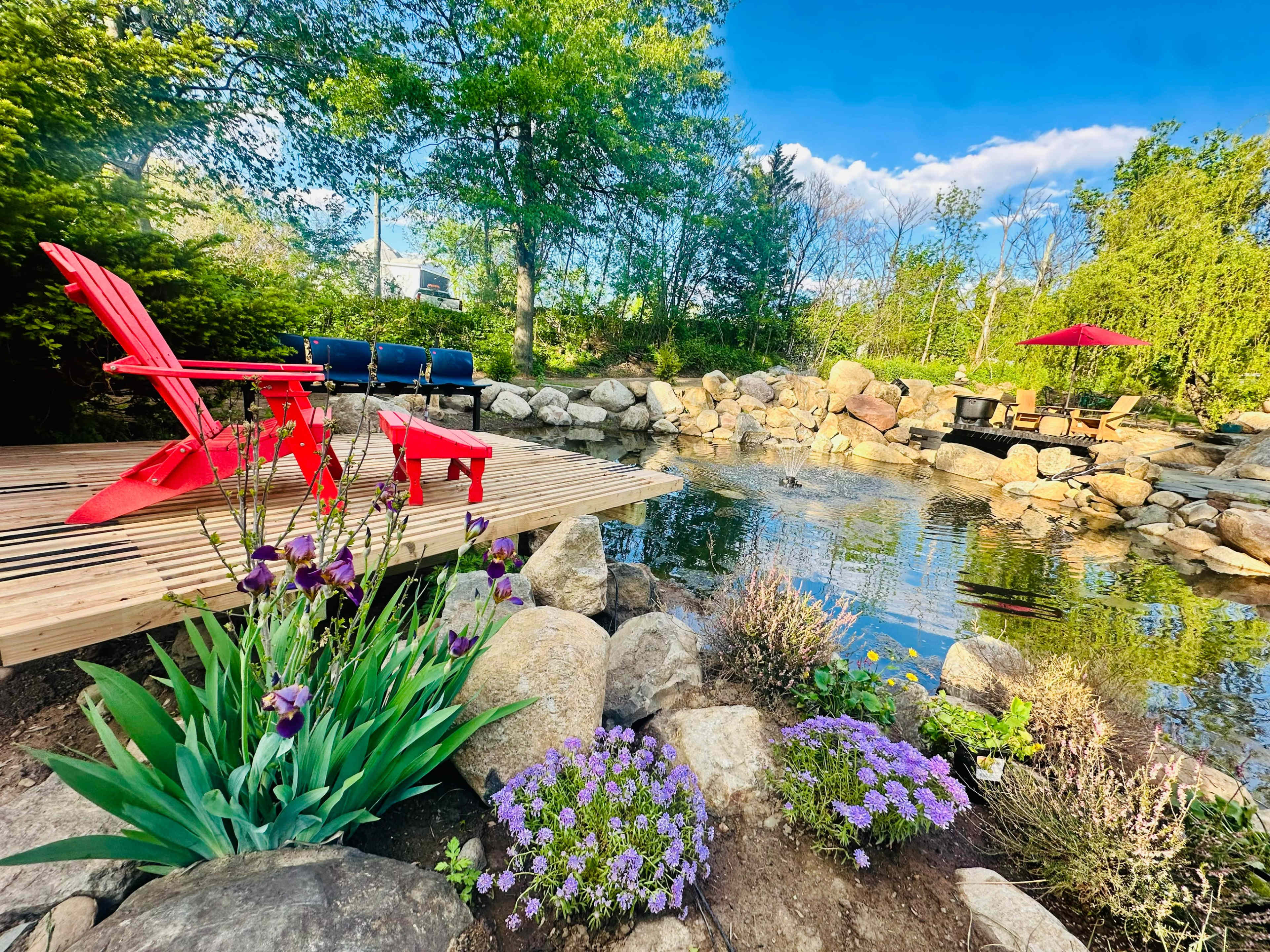 A wooden deck with red chairs overlooks a pond surrounded by rocks and a variety of flowering plants.