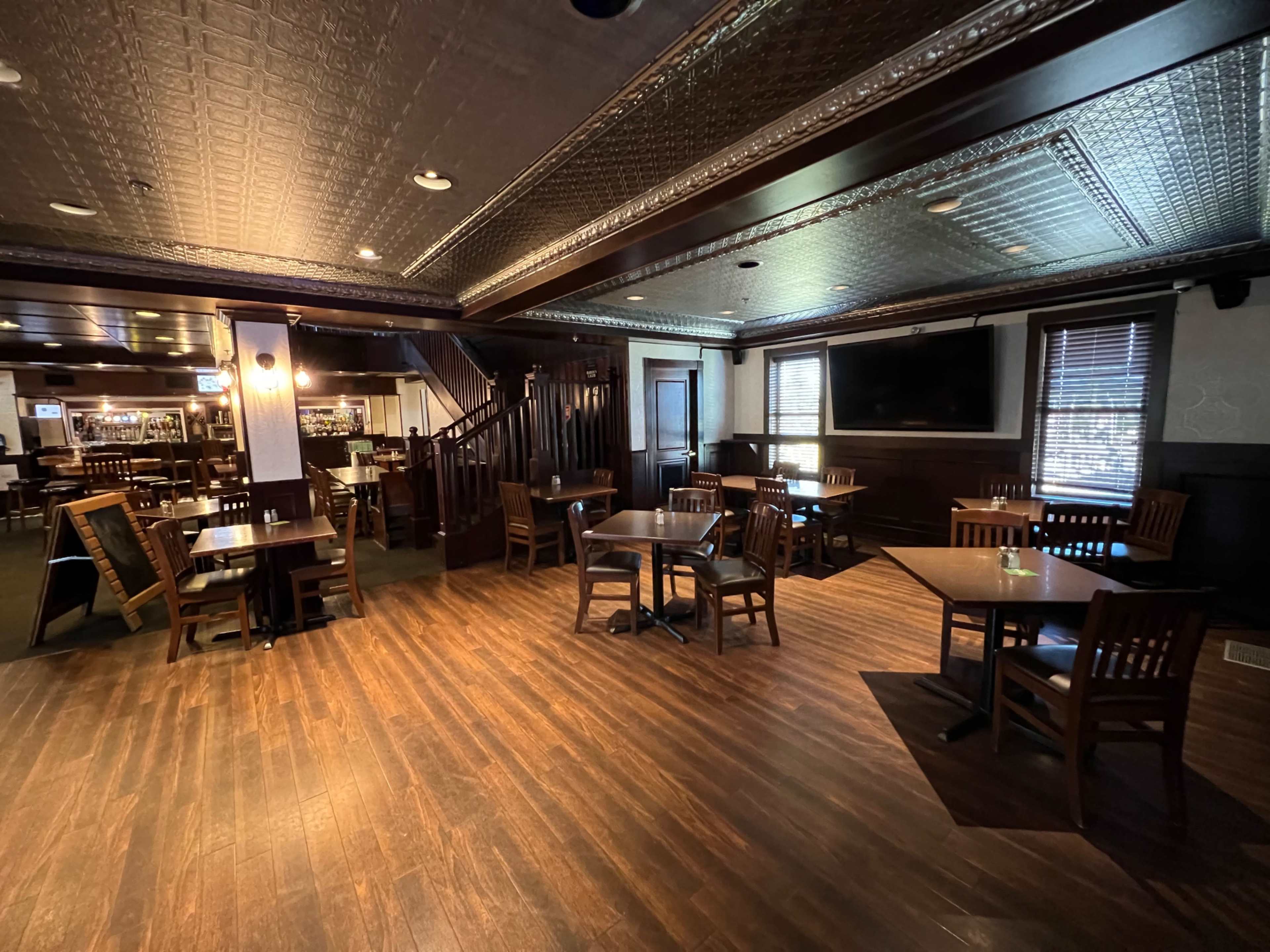 An interior view of a dimly lit restaurant with wooden tables and chairs arranged on a hardwood floor, featuring a staircase and a bar in the background.