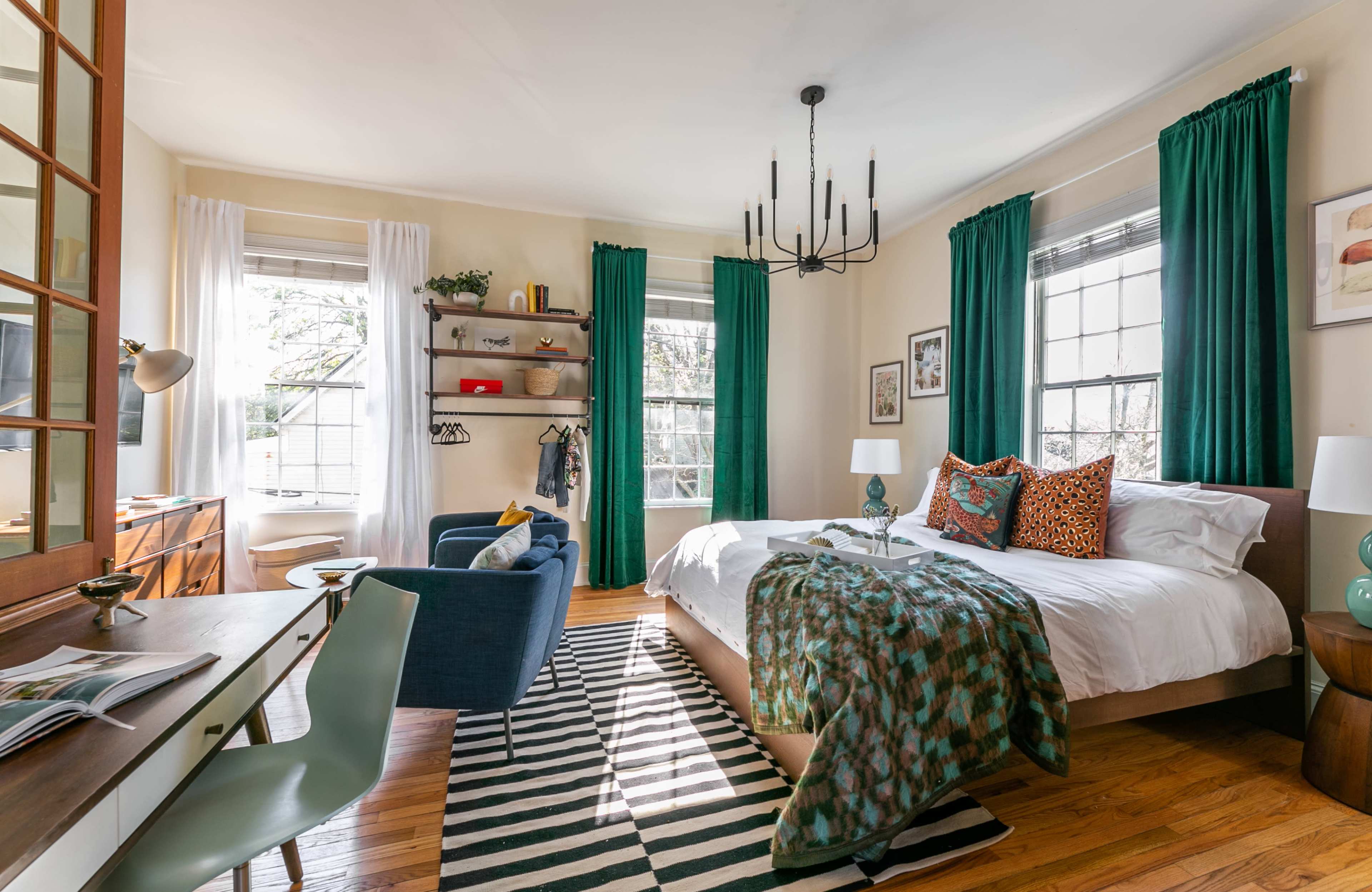A bright bedroom featuring a bed with patterned pillows, a desk, a chair, and large windows draped with green curtains.