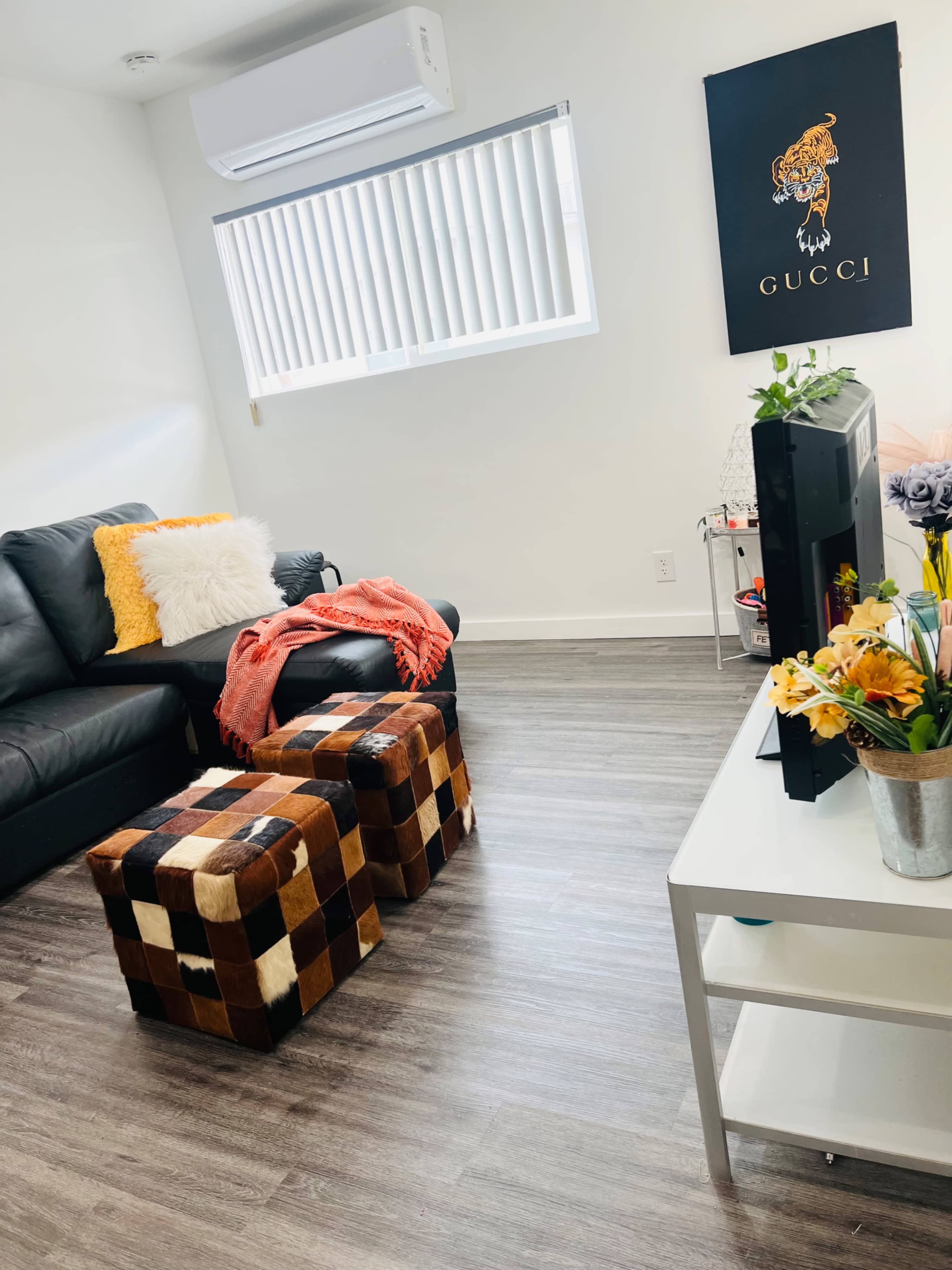 A cozy living room with a black leather couch, two patterned ottomans, a white coffee table with flowers, and a wall featuring a Gucci art piece.