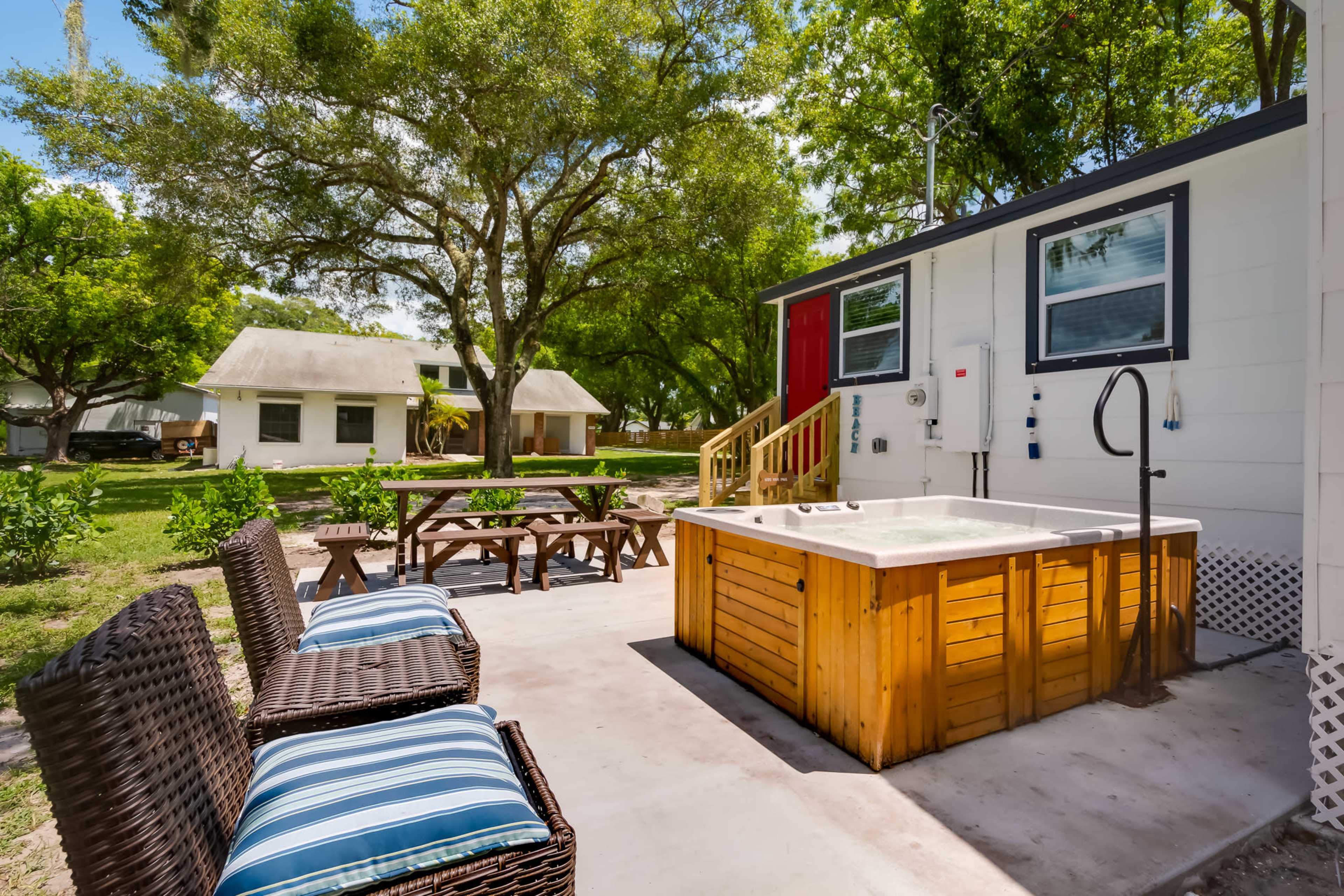 A hot tub is positioned on a patio next to a small seating area and a white building with a red door, surrounded by trees and another building in the background.