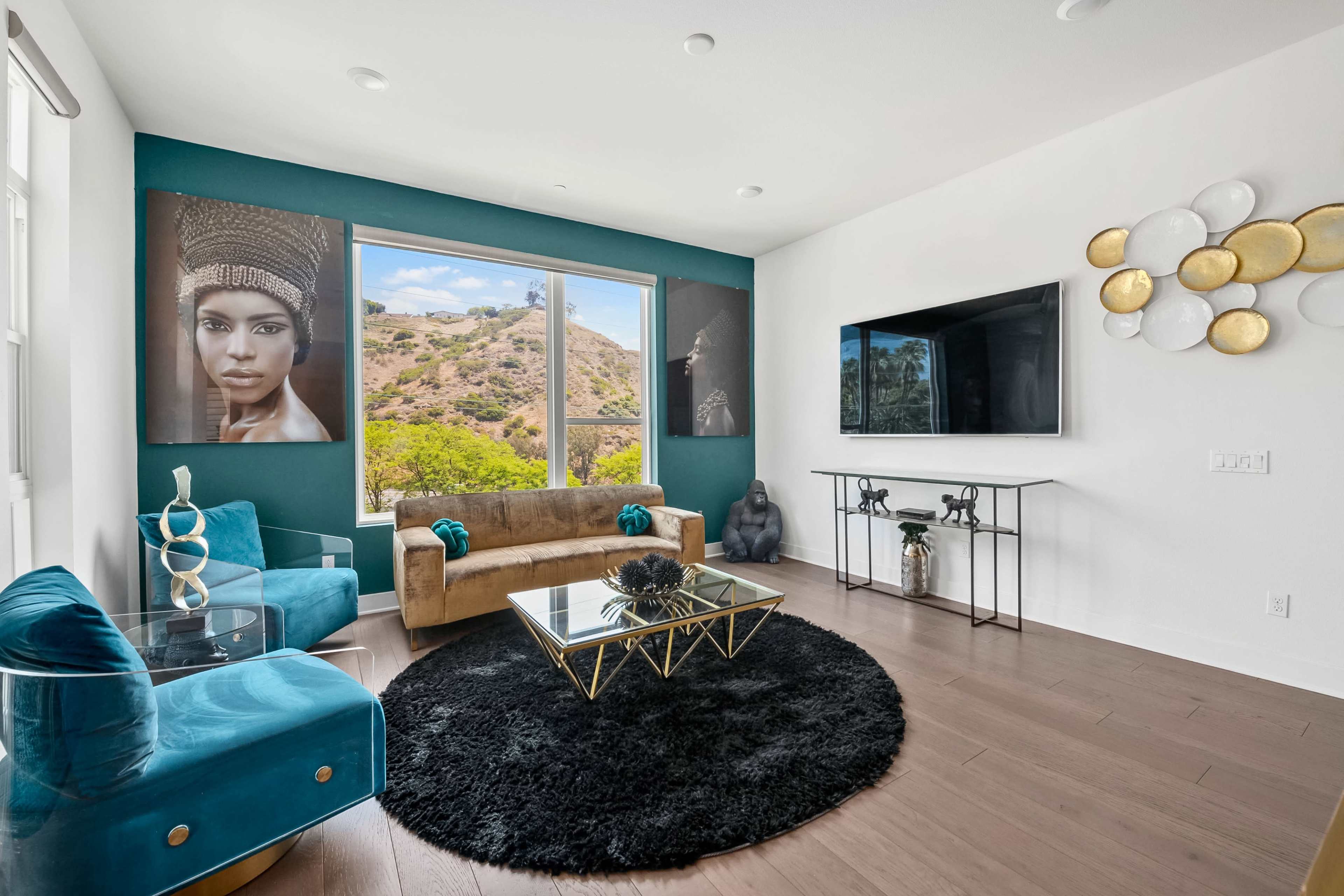 The living room features a modern design with a gold sofa, turquoise chairs, a black area rug, and large windows displaying a hillside view.