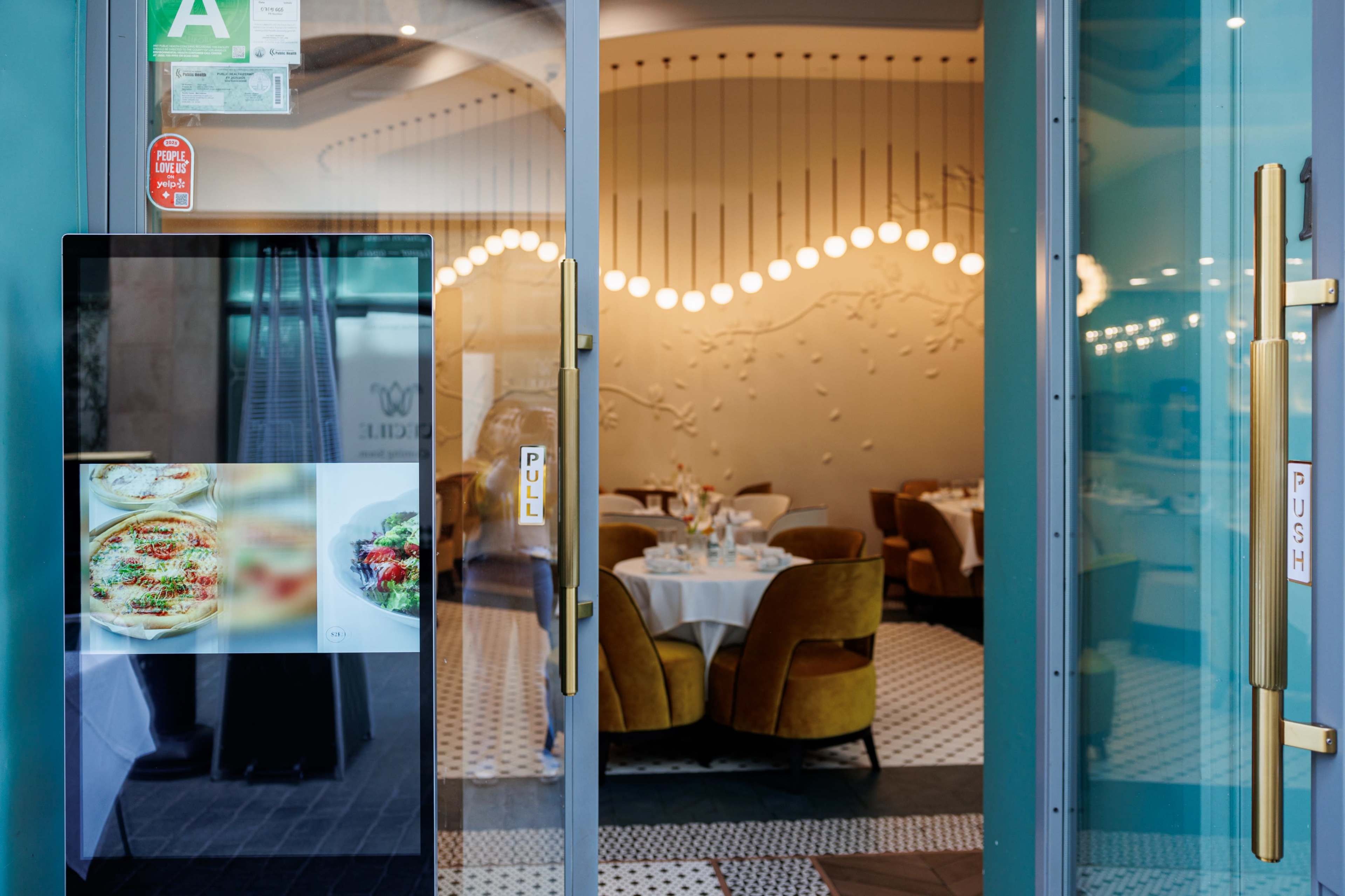 The image shows the entrance of a restaurant featuring glass doors with push and pull signs, a digital menu display, and a dining area visible in the background.