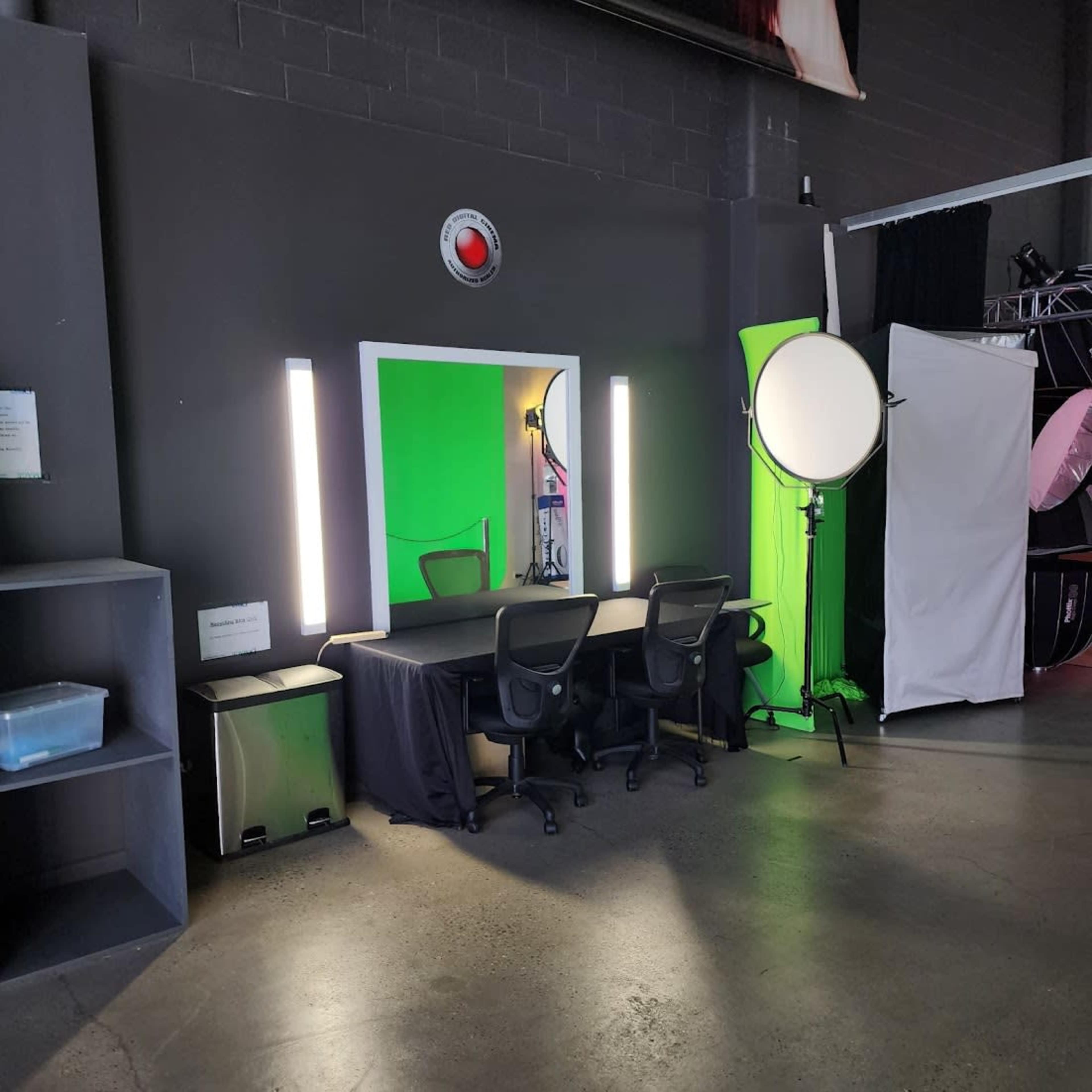 The image shows a photo studio setup with a green screen, two bright lights, and three chairs arranged at a table.