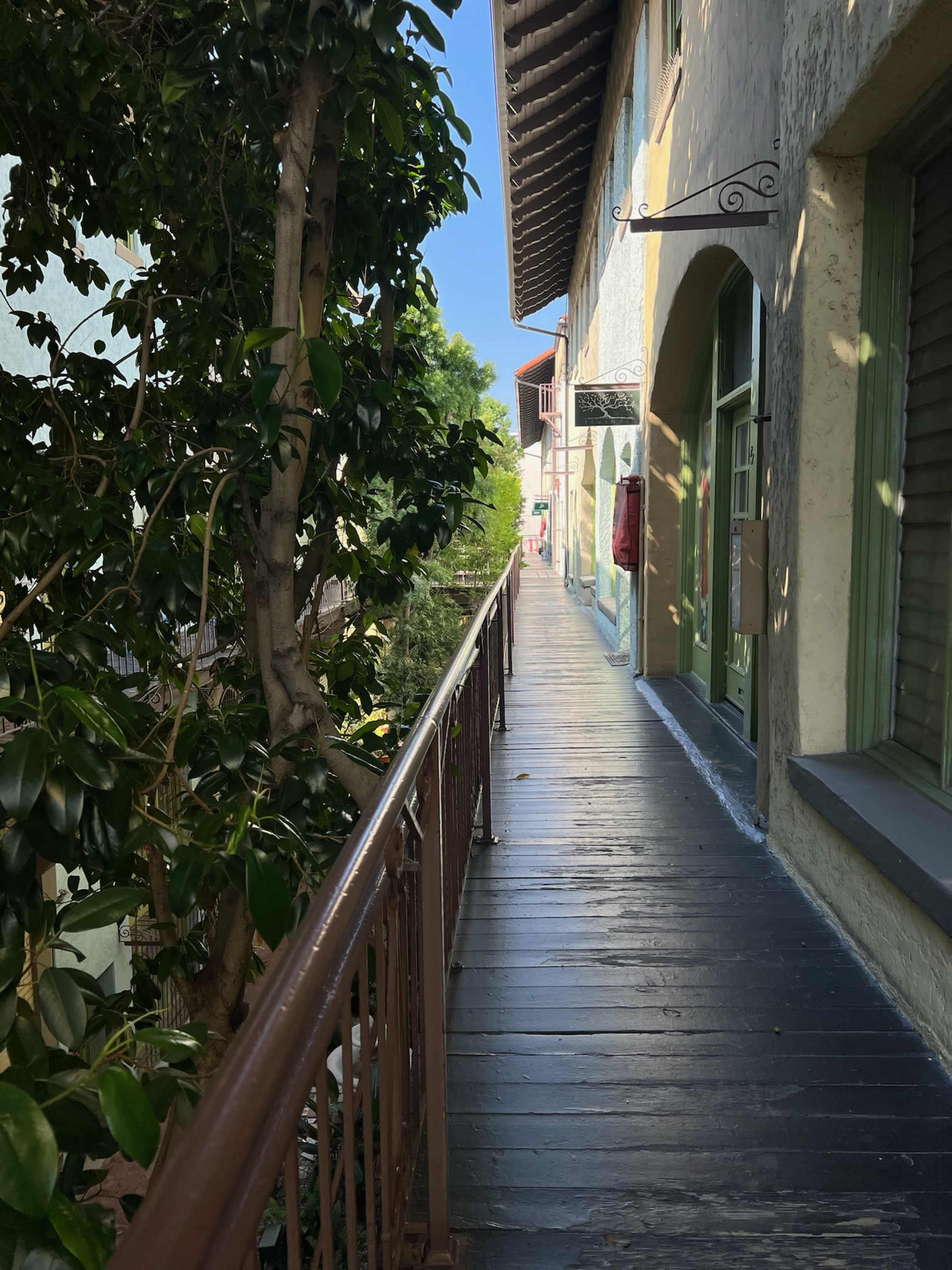 A narrow wooden walkway enclosed by buildings and greenery under a clear blue sky.