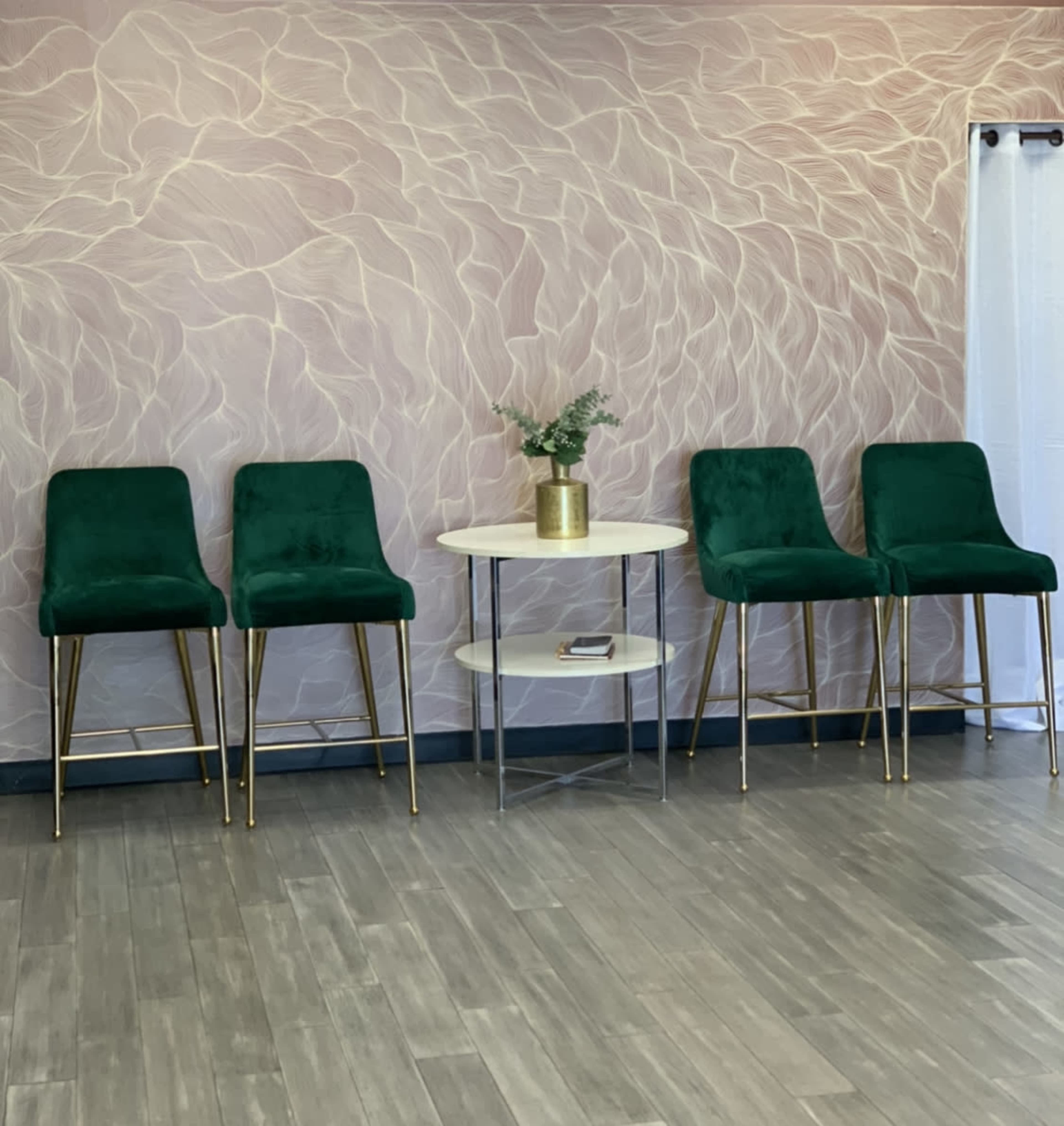 Four green velvet bar stools with gold legs arranged around a small round table on a wooden floor against a patterned wall.