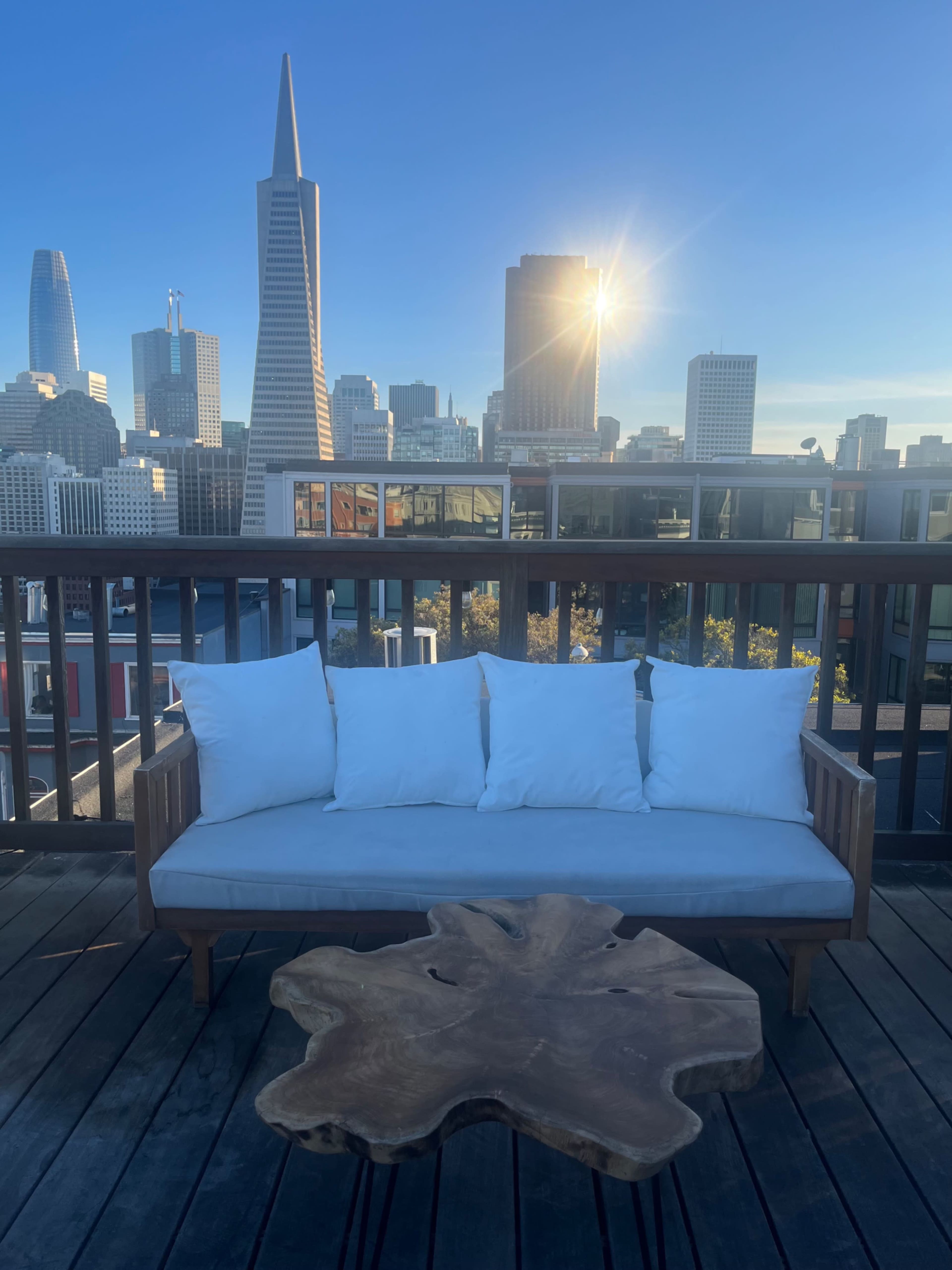 A wooden couch with white cushions and a natural wood coffee table is positioned on a balcony overlooking a city skyline at sunset.