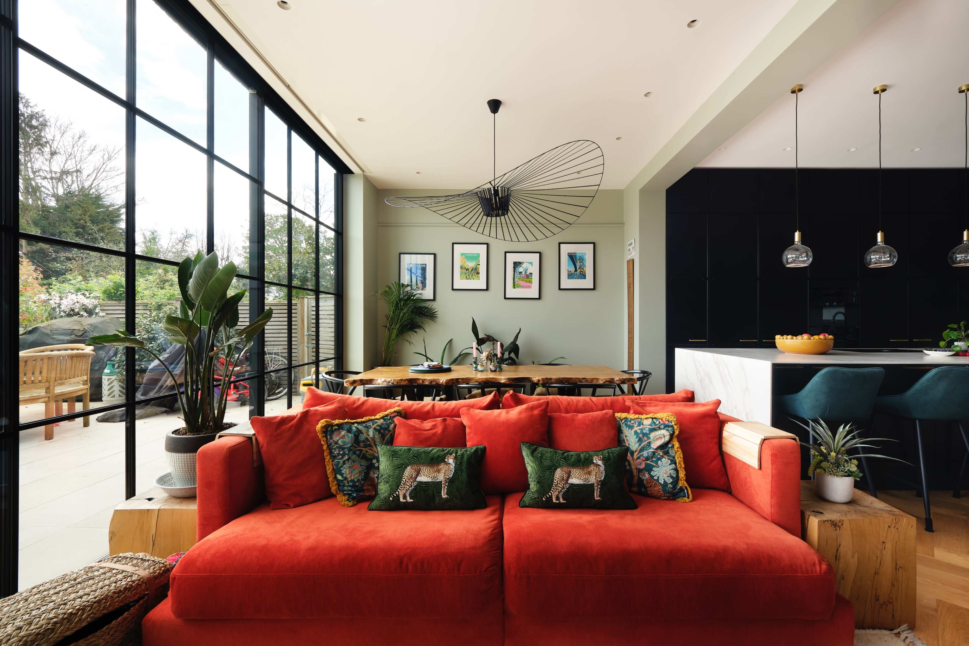 A bright living room features a large red sofa with decorative pillows, large windows, and a dining area adjacent to a modern kitchen.