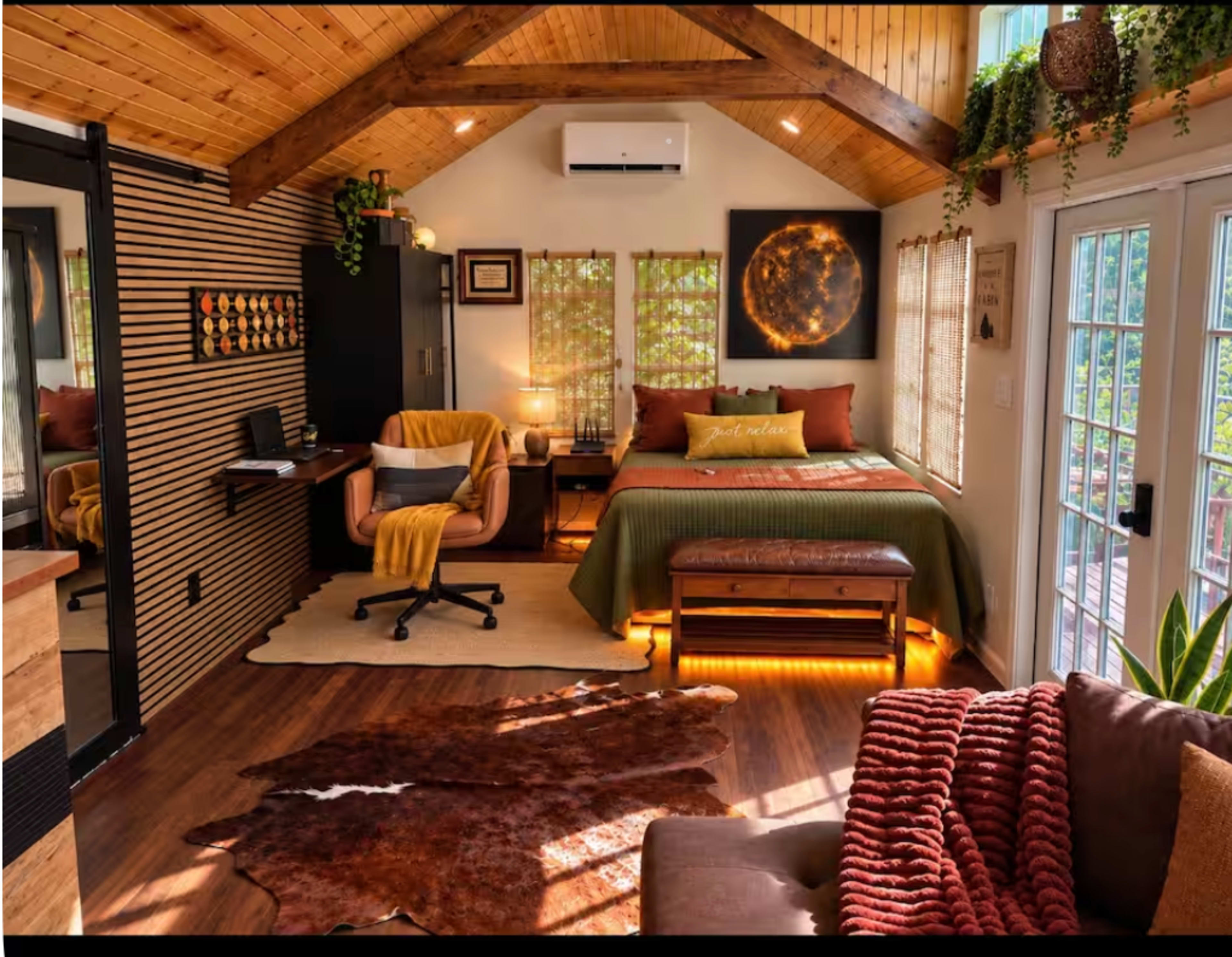 The image shows a cozy, well-lit bedroom with a wooden ceiling, a bed with green bedding, a sitting area, and large windows letting in natural light.