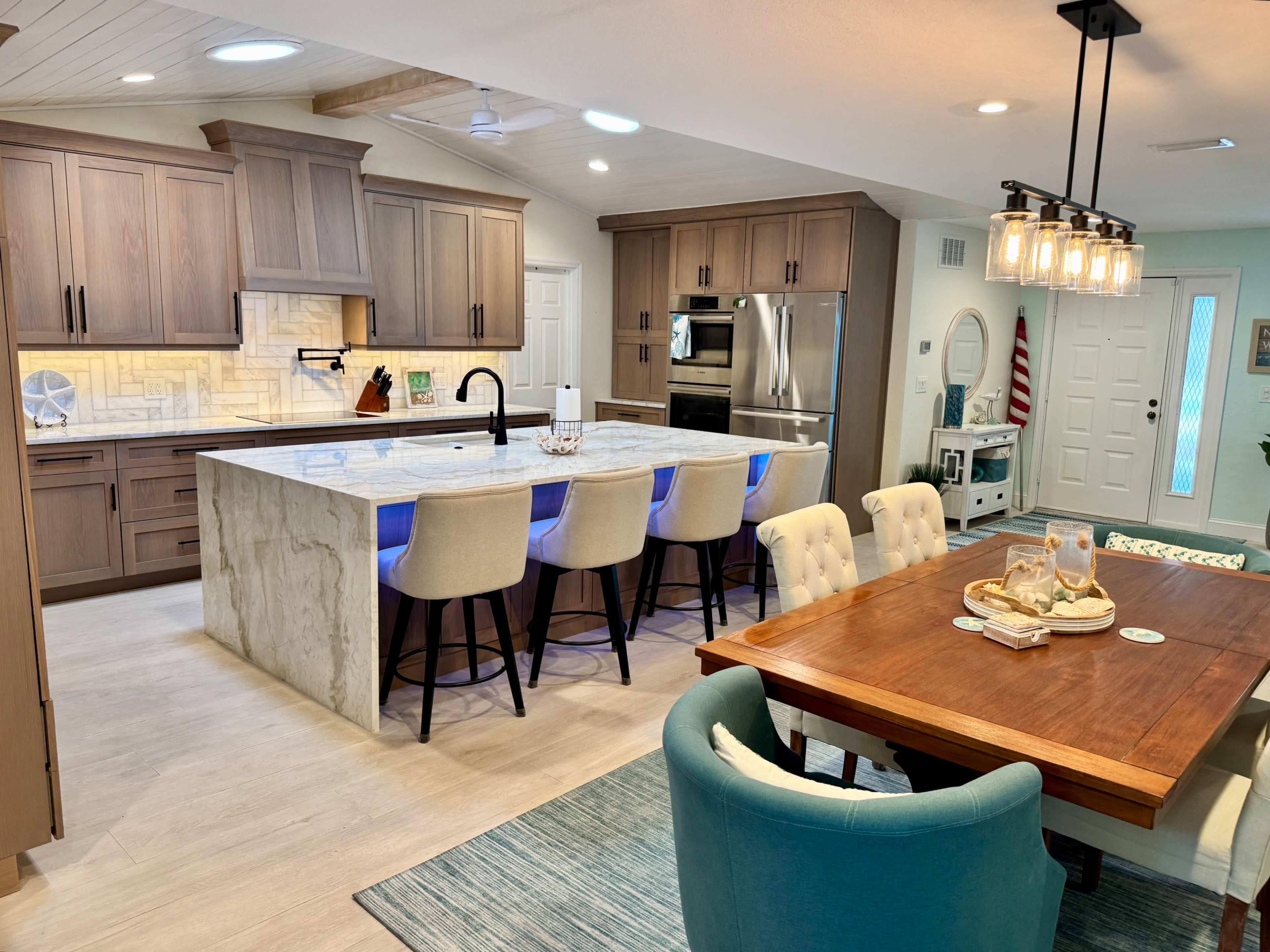 The image shows a modern kitchen with wooden cabinets, a large marble island with blue LED lighting, and an adjacent dining area featuring a wooden table and upholstered chairs.