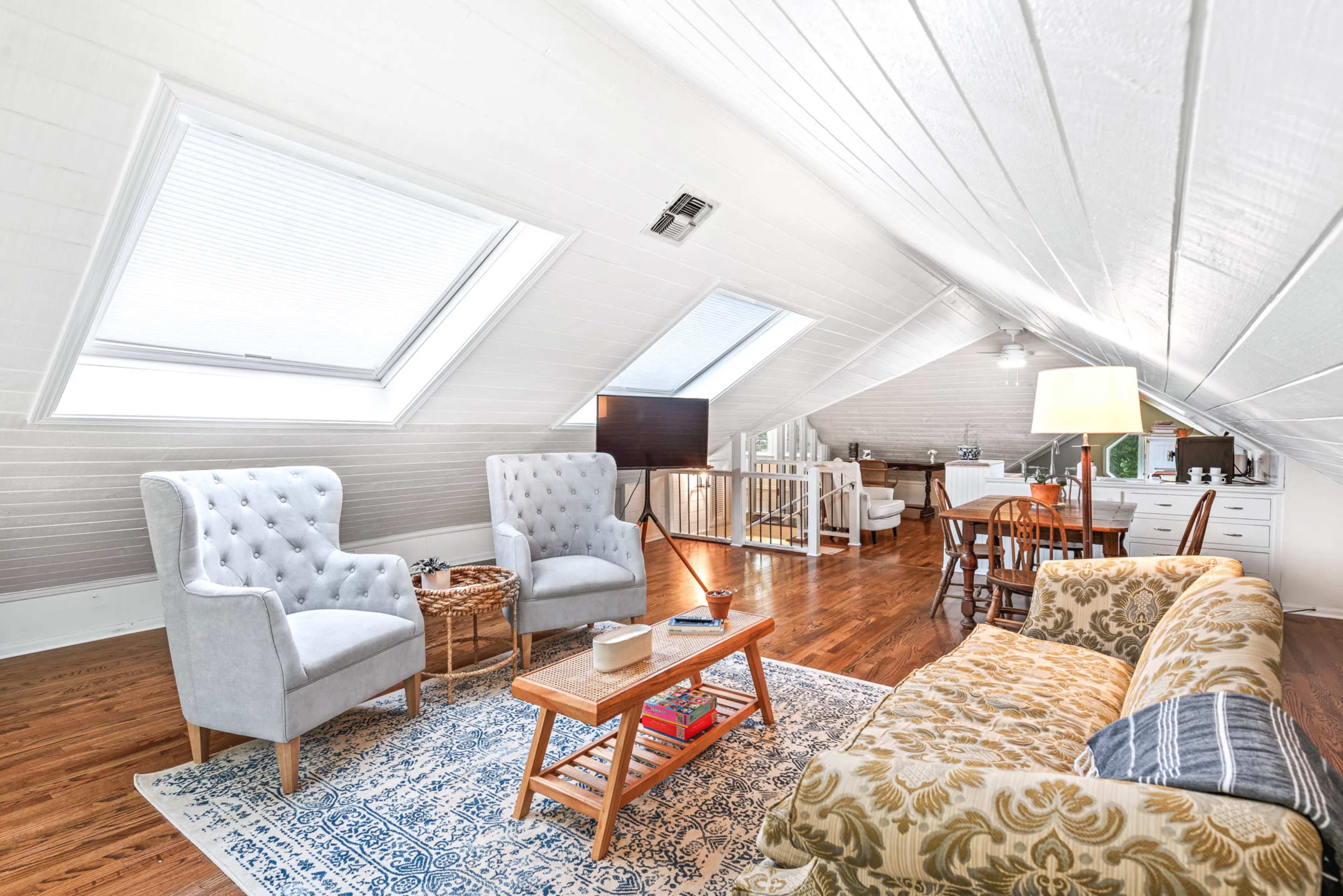 The image shows a bright attic living space with two upholstered chairs, a sofa, a coffee table, and a dining area in the background.