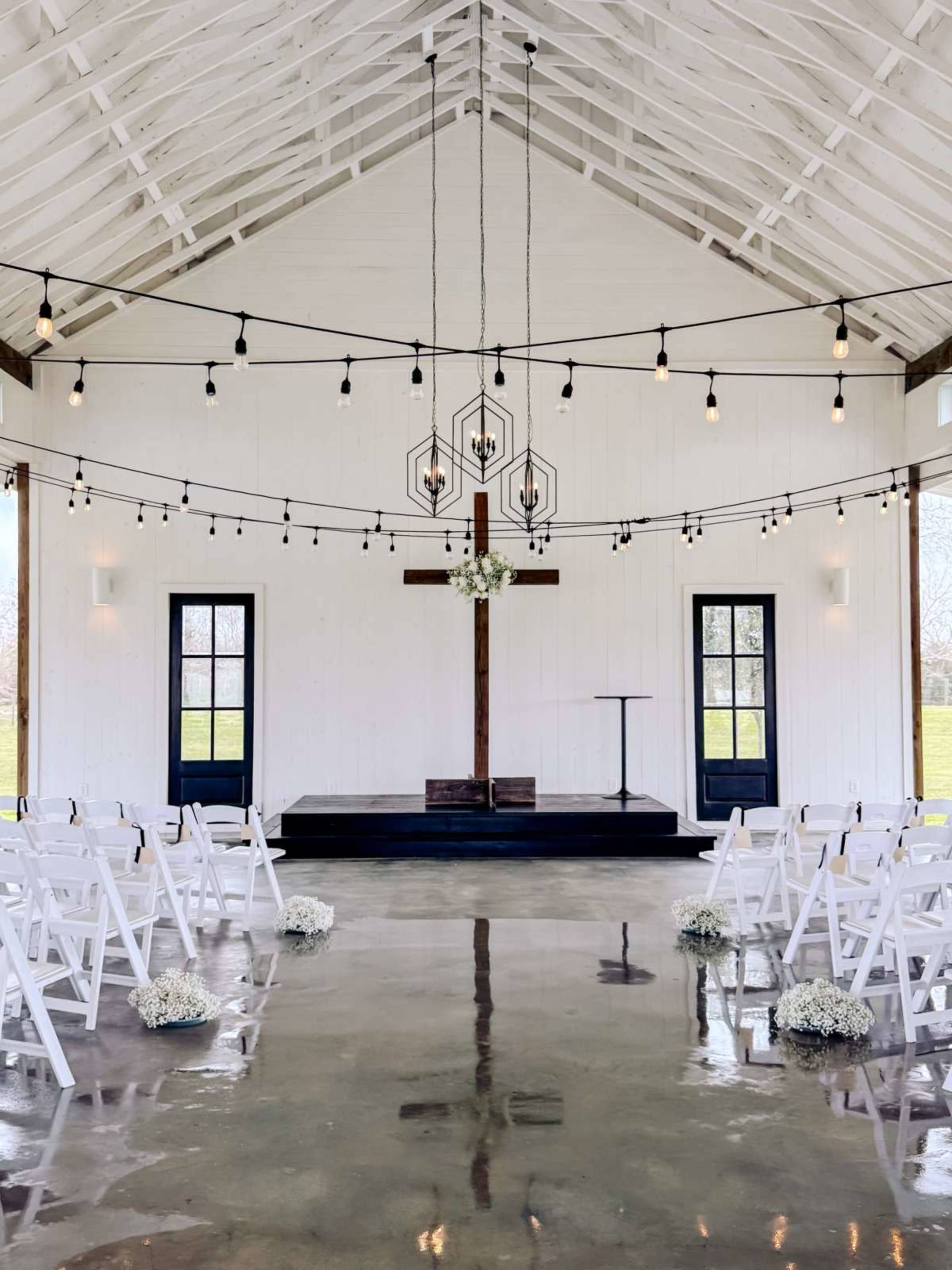 The image shows a wedding ceremony setup inside a spacious venue featuring white chairs arranged in rows, a wooden cross adorned with flowers, and decorative light strands hanging from the ceiling.