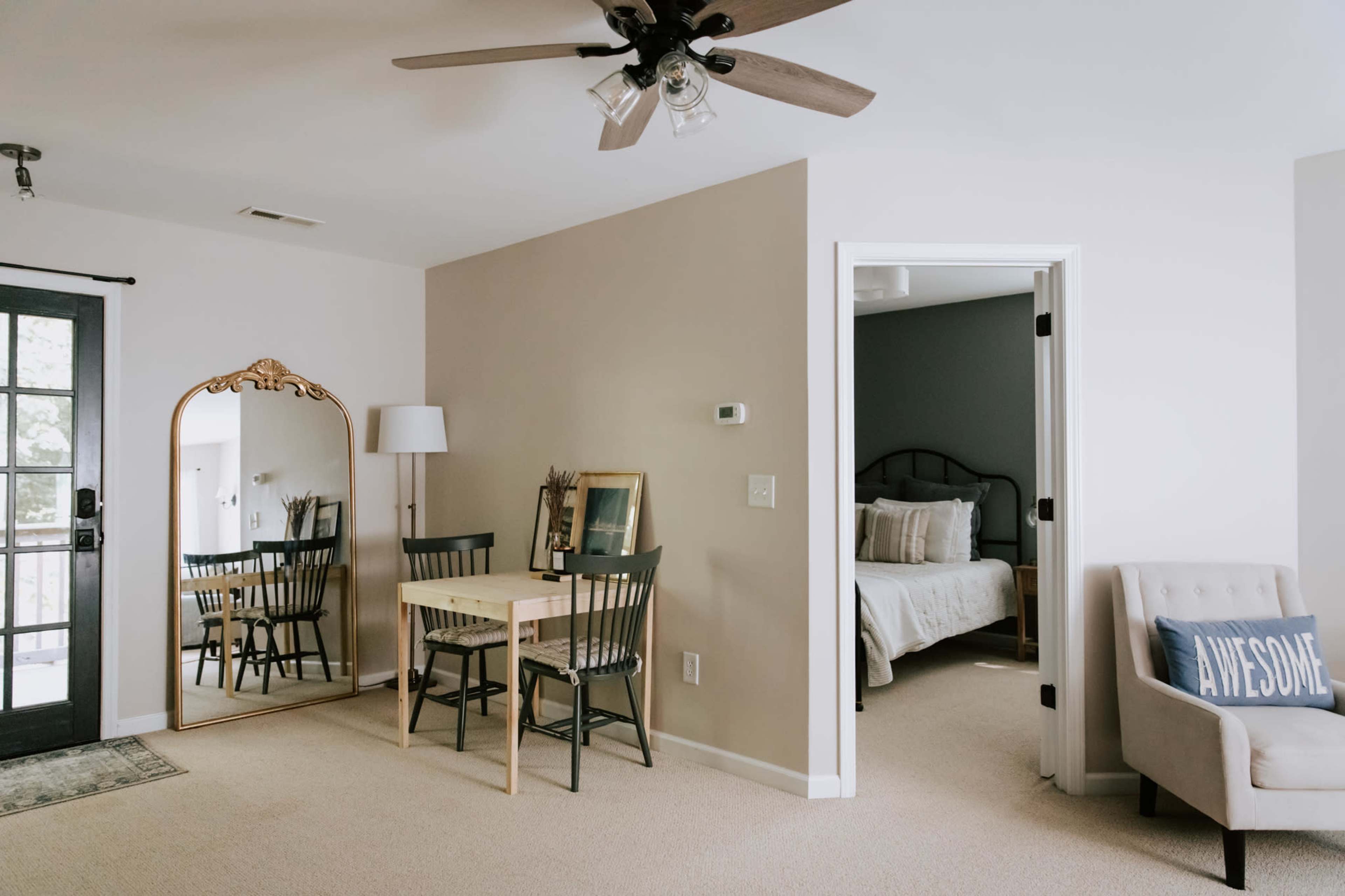 A small room features a dining area with a table and two chairs, a mirror, a couch, and a doorway leading to a bedroom.