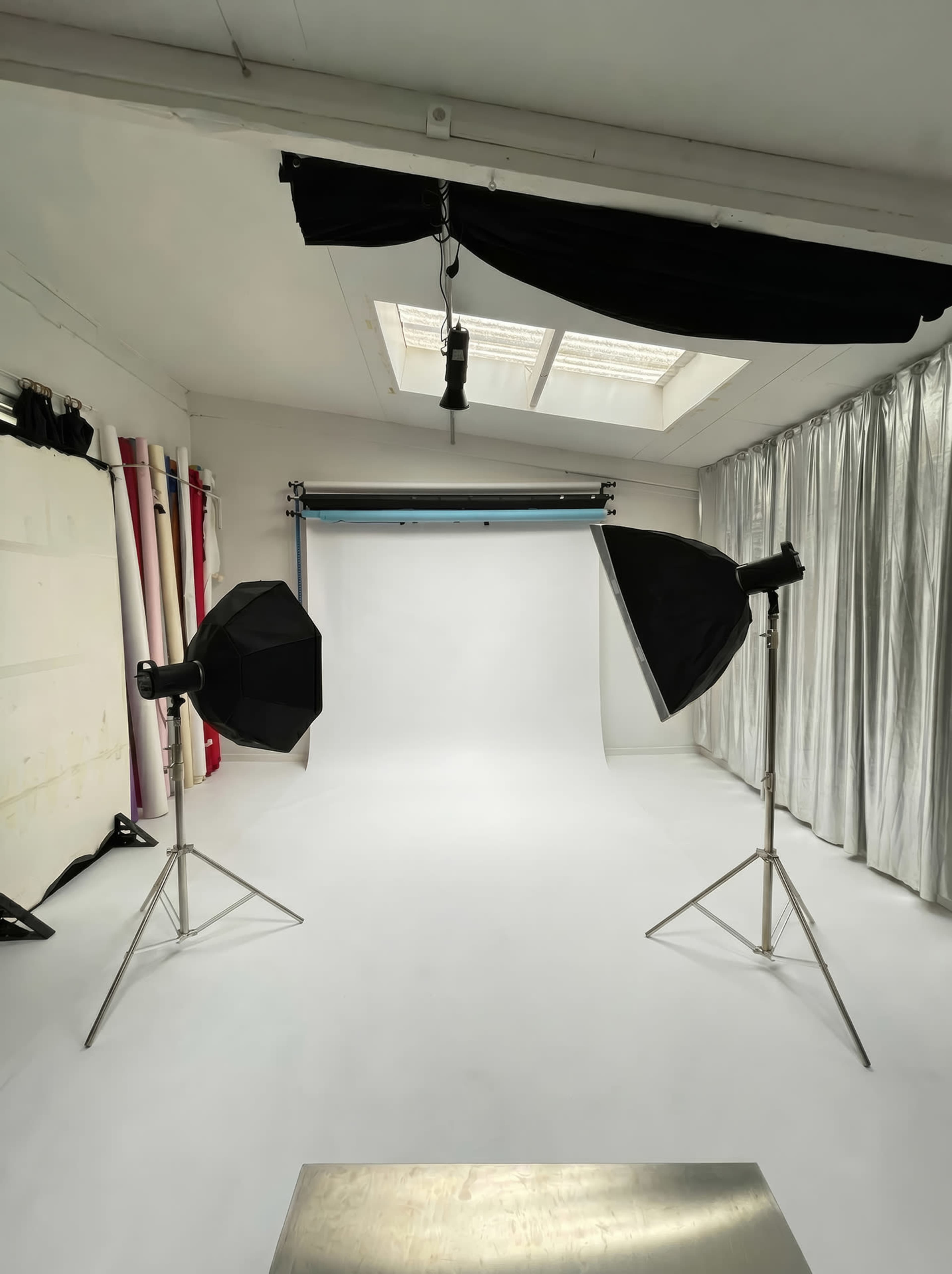 The image shows a photography studio with two softbox lights, a blank backdrop, and various drapes along the walls.
