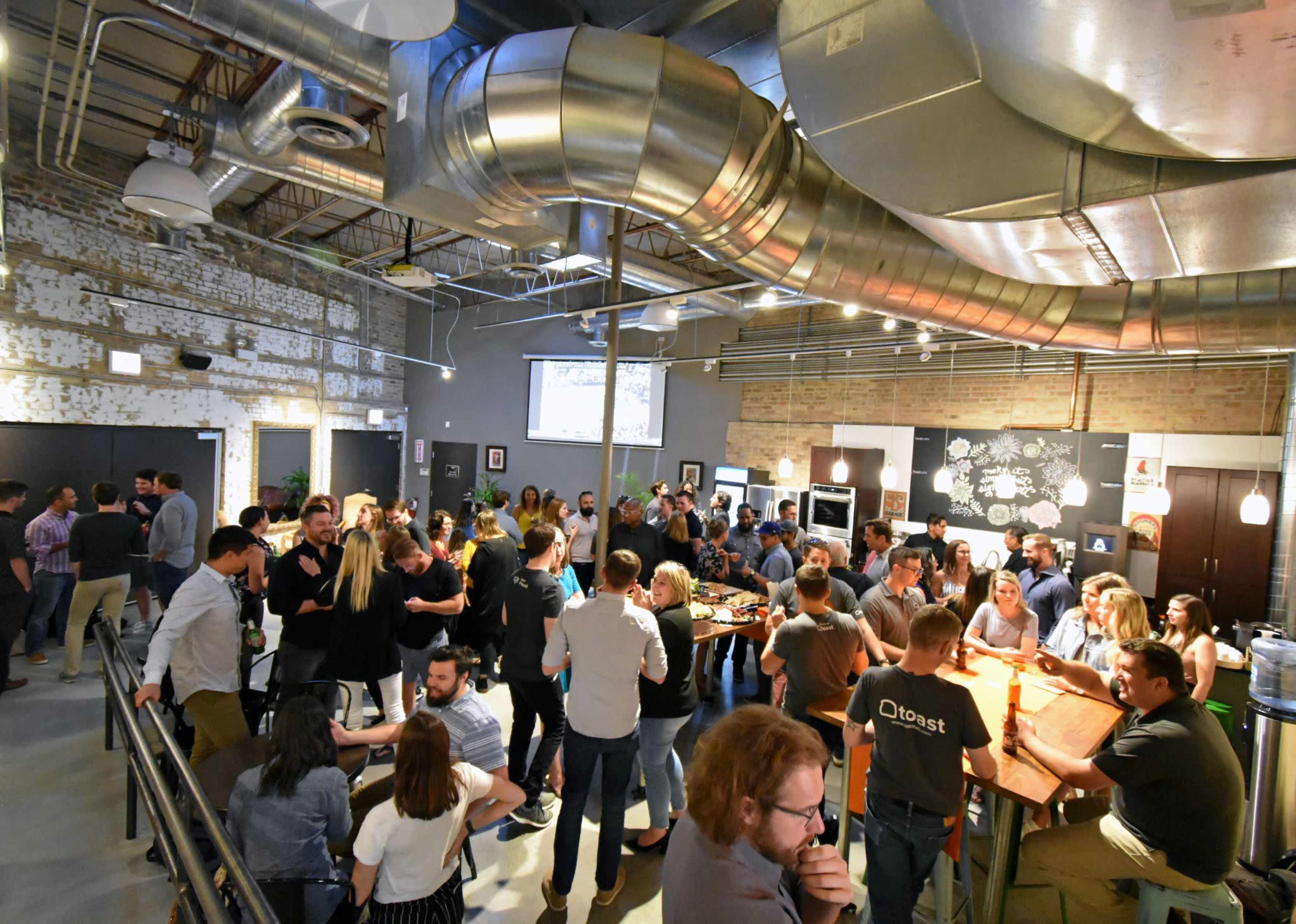 A large group of people gathers socializing in a spacious, well-lit industrial-style room with exposed ducts and a modern kitchen area.