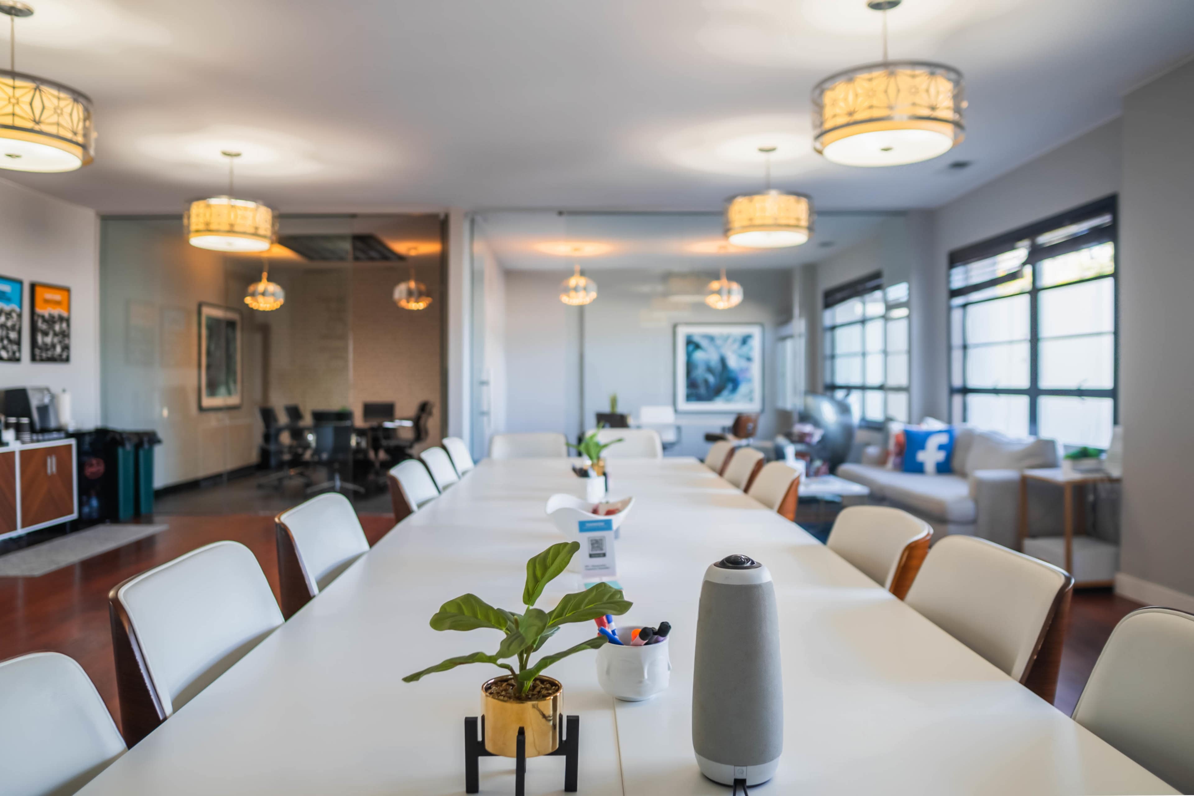 A long, white conference table is set in a well-lit office with modern lighting and decor, featuring a small plant and various office supplies.