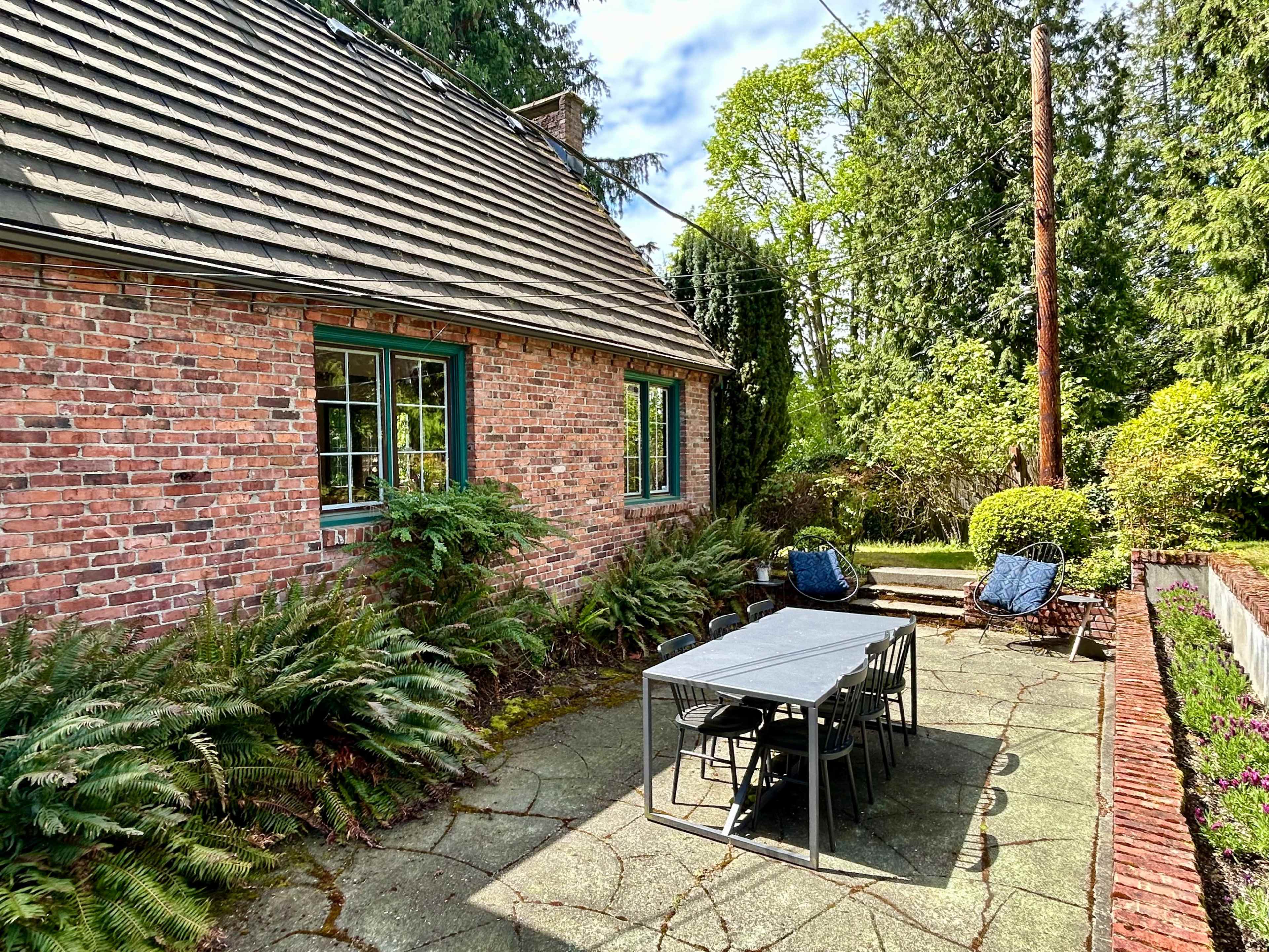 A brick house with a patio area featuring a dining table and two chairs, surrounded by greenery.