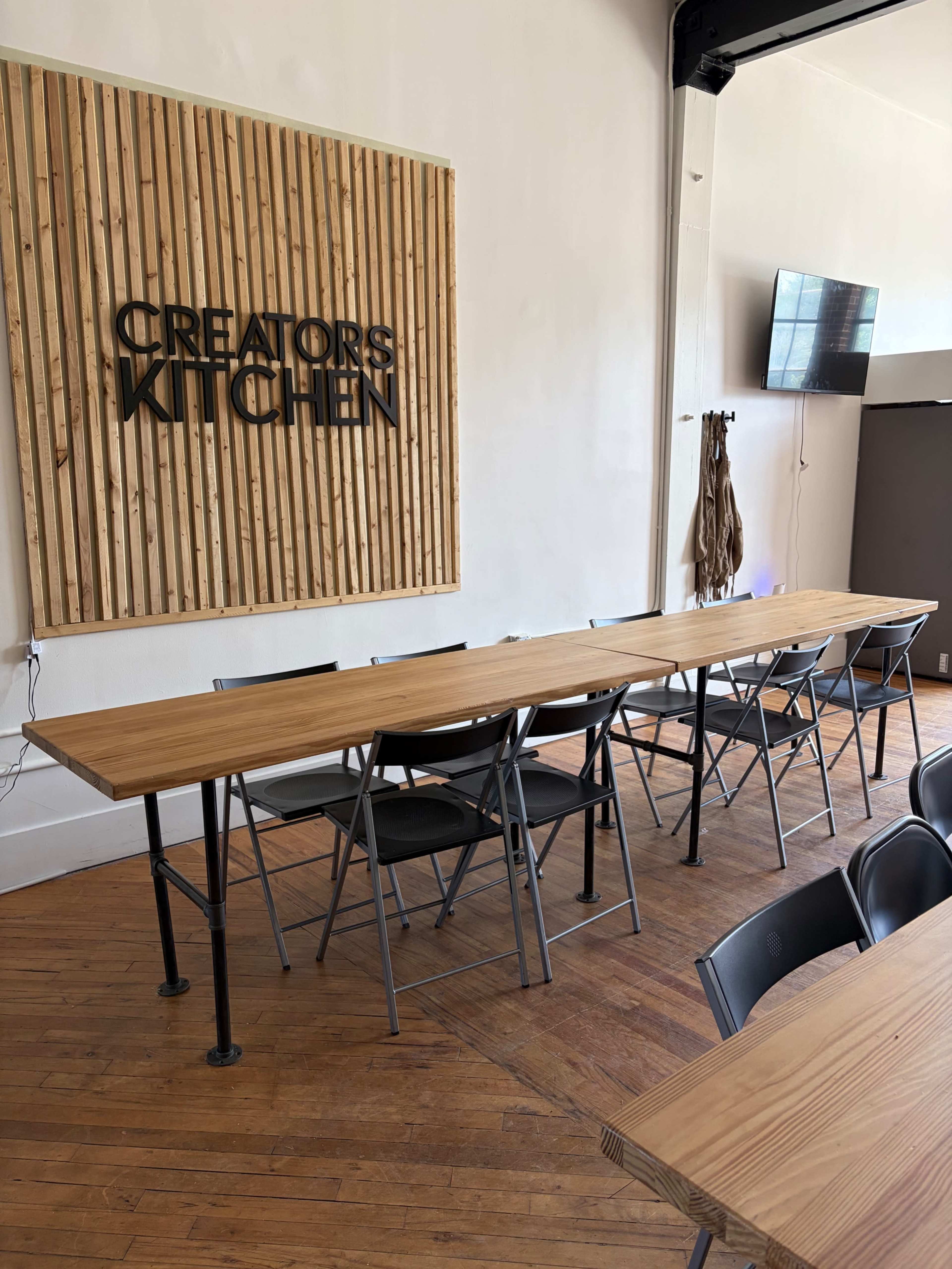 A long wooden table with black folding chairs is set in a room featuring a wooden wall sign that reads "CREATORS KITCHEN."